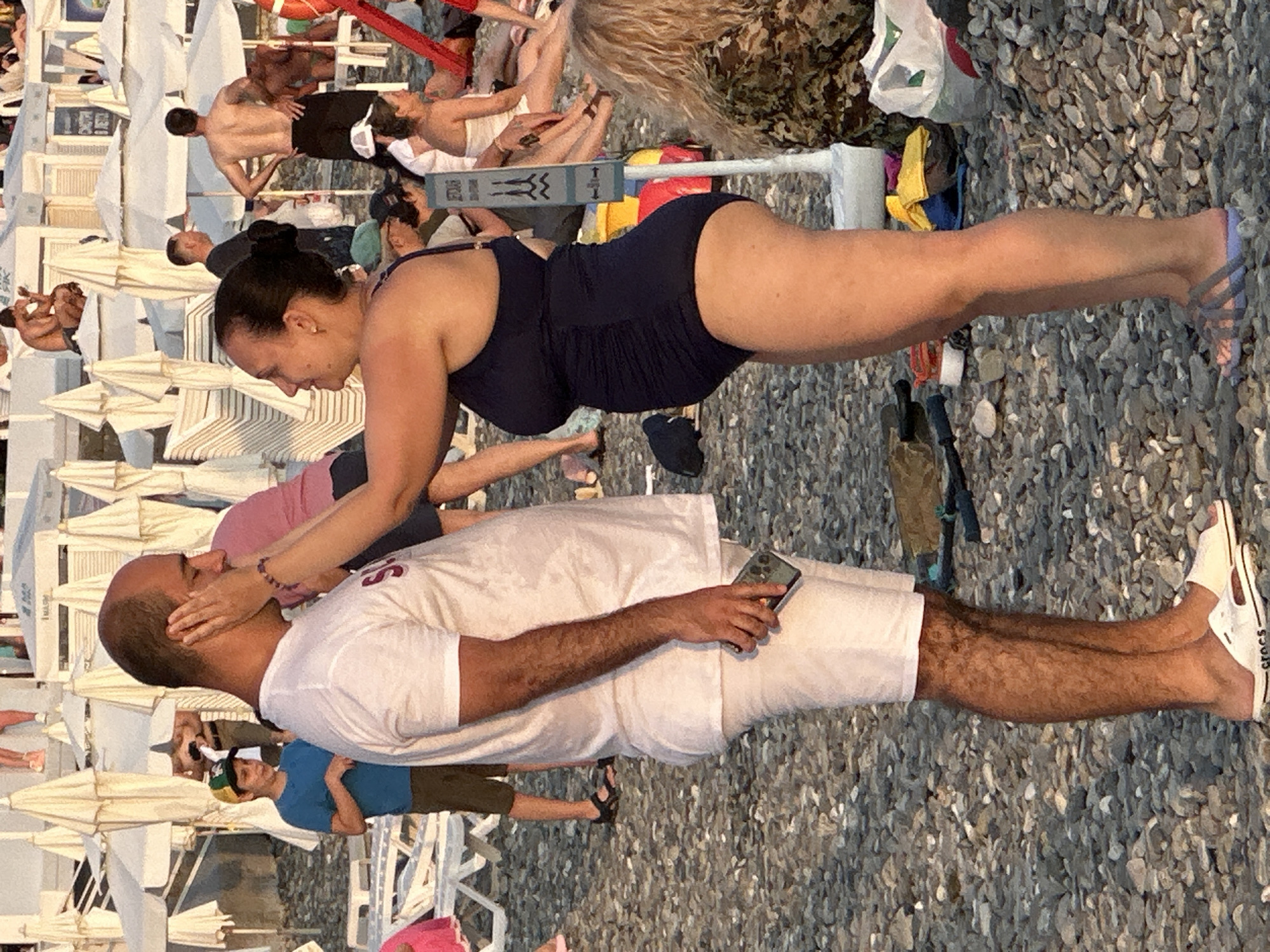 The image depicts a group of people at a beach in Sochi, Russia. The sun appears to be setting, casting a warm glow on the scene. The beach is covered in pebbles, and there are numerous beach chairs and umbrellas set up.
In the foreground, a man and a woman are interacting. The man, wearing a white t-shirt and white shorts, is lying on the ground with his eyes closed, appearing relaxed. The woman, in a dark blue swimsuit, is leaning over him, her hands on his face. She has dark hair and is wearing earrings.
Further back, other individuals are visible, some sitting on chairs and others walking around. One person is wearing a blue t-shirt and a green and yellow cap. The overall atmosphere seems casual and leisurely, consistent with a beach setting. The presence of beach chairs and umbrellas suggests a public beach area.