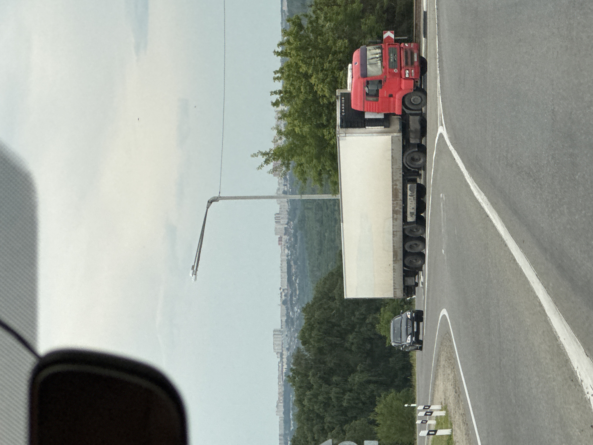 A red truck with a white trailer is parked on the side of a road in Mikhaylovsk, Russia. The truck appears to be a semi-trailer truck. The trailer is large and white, extending behind the truck cab. A black car is visible behind the trailer, also parked on the roadside. The road has white lane markings and a grey asphalt surface.
In the background, there are lush green trees and a distant cityscape with buildings. A tall pole with a light fixture is also present near the truck. The sky is overcast and appears to be daytime. The overall scene suggests a stop or rest area along a highway. No people are clearly visible in the image. The word "CARRIER" is visible on the side of the trailer.
