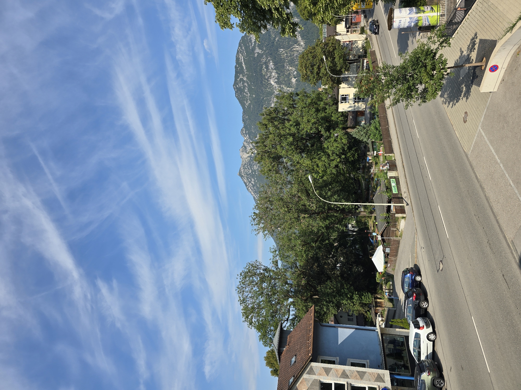 The image depicts a street scene in Garmisch-Partenkirchen, Germany, under a bright blue sky with wispy white clouds. In the foreground, a paved road runs diagonally across the frame, with parked cars lining the left side. Several residential buildings with tiled roofs are visible, one prominently featuring a blue facade. Beyond the buildings, a dense line of mature green trees forms a backdrop, obscuring a playground area and some smaller structures. In the distance, a majestic mountain range with rocky outcrops and patches of vegetation dominates the horizon. A single black car is in motion on the road in the far right of the image, and a green utility pole with advertisements stands on the sidewalk. The overall impression is one of a tranquil summer day in a picturesque Bavarian town.