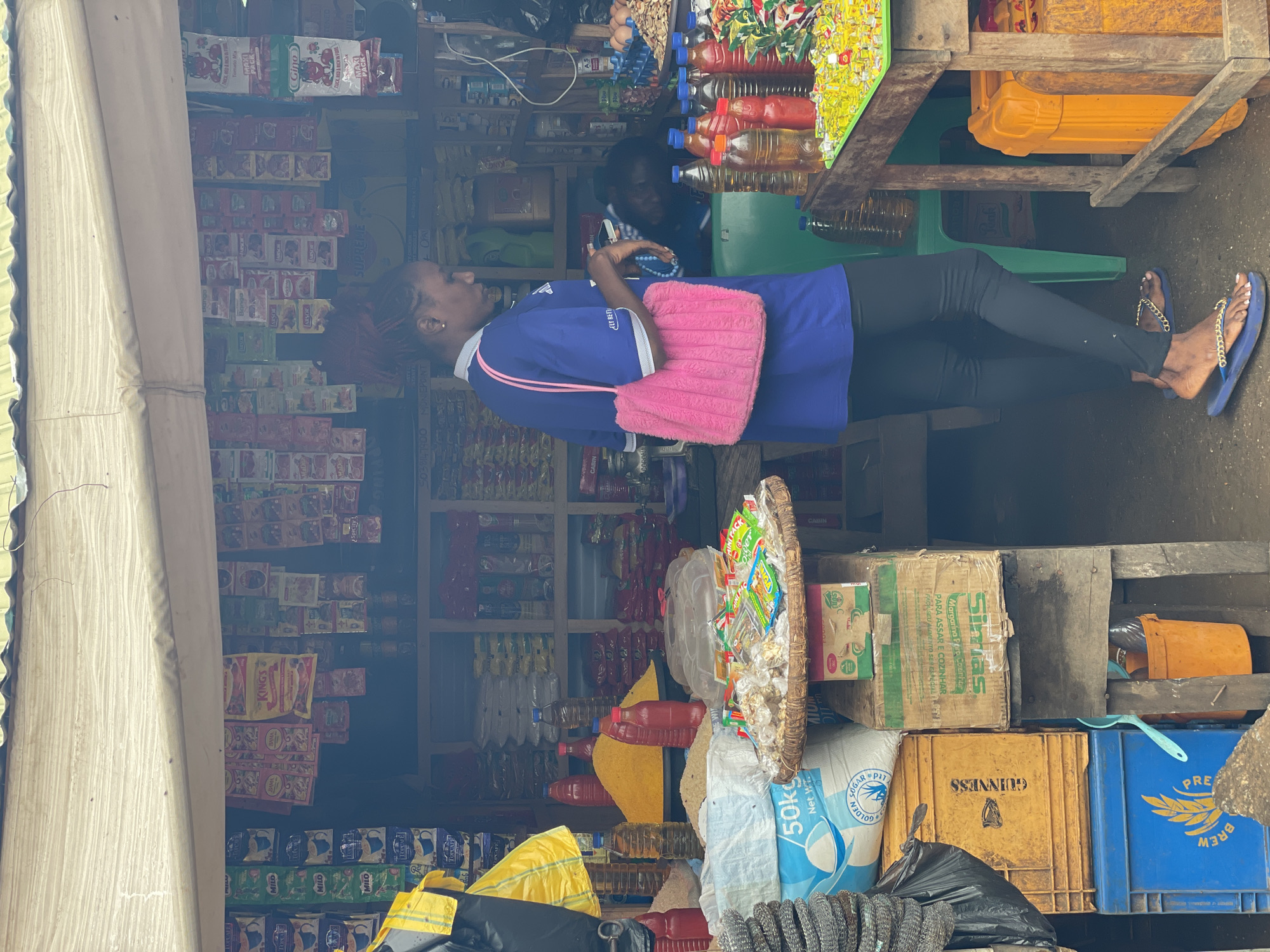 A young woman with red-streaked hair, wearing a blue shirt and black leggings, stands in a market stall. She is holding a pink, fluffy purse. The stall is filled with various goods, including bags of sugar, bottles of oil and sauce, and numerous packets of food products hanging from the ceiling. In the background, other people can be seen. The scene is located in the city of Akwanga, Nigeria. The market stall appears to be an open-air setup with a corrugated metal roof covered by a white tarp. The ground is dirt. Various crates and containers, some yellow and blue, are stacked around the stall.