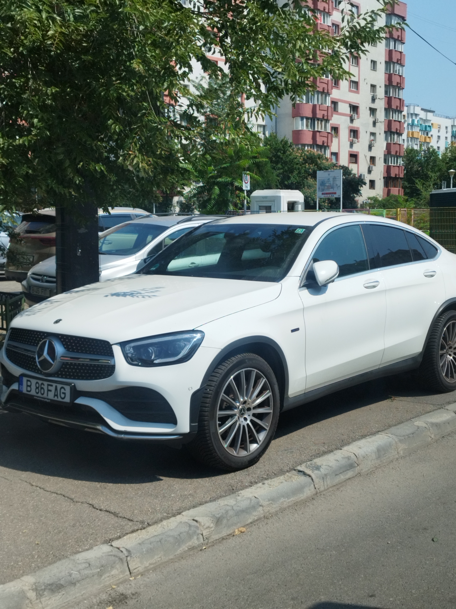A white Mercedes-Benz GLC Coupe is parked on a paved street in front of a residential building in Bucharest, Romania. The car has a license plate that reads "B 86 FAG". A large tree with green leaves is in the foreground, casting shadows on the car and the street. The building in the background is tall and appears to be made of concrete with pink and white accents. There are several windows on the building, and some of them have balconies. The sky is blue and clear, indicating it is a sunny day. The street is lined with a concrete curb. Other cars are visible parked behind the Mercedes.
