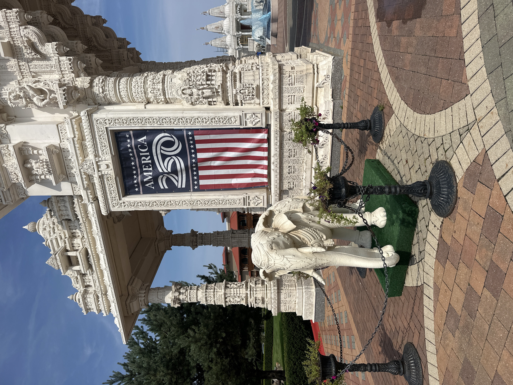 The image shows the exterior of a Hindu temple in Bartlett, Illinois. In the foreground, a white elephant statue stands on a green platform, surrounded by black metal posts and chains. Behind the statue, a large banner hangs on the temple wall, displaying "AMERICA 250" in white text over a background of the American flag. The temple itself is ornate, constructed of white stone with intricate carvings. In the background, other temple structures and a blue fountain are visible. The ground is paved with bricks in various shades of brown and gray. The sky is clear and blue.