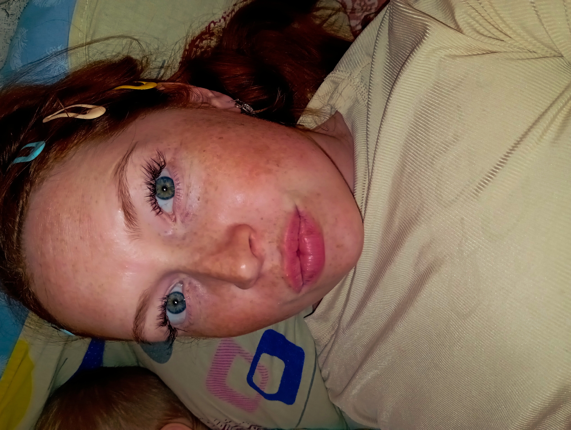 A close-up, tilted, indoor shot shows a woman with light skin and many freckles, lying down. Her hair is predominantly red-brown, with hair clips in blue, yellow, and white visible. She has blue eyes. She is wearing a light-colored ribbed shirt. The background includes a patterned pillow in blue, white, pink and a small section of a baby's head is in the lower left corner.
I cannot tell the location in the city of Yurgamysh, Russia from this image.