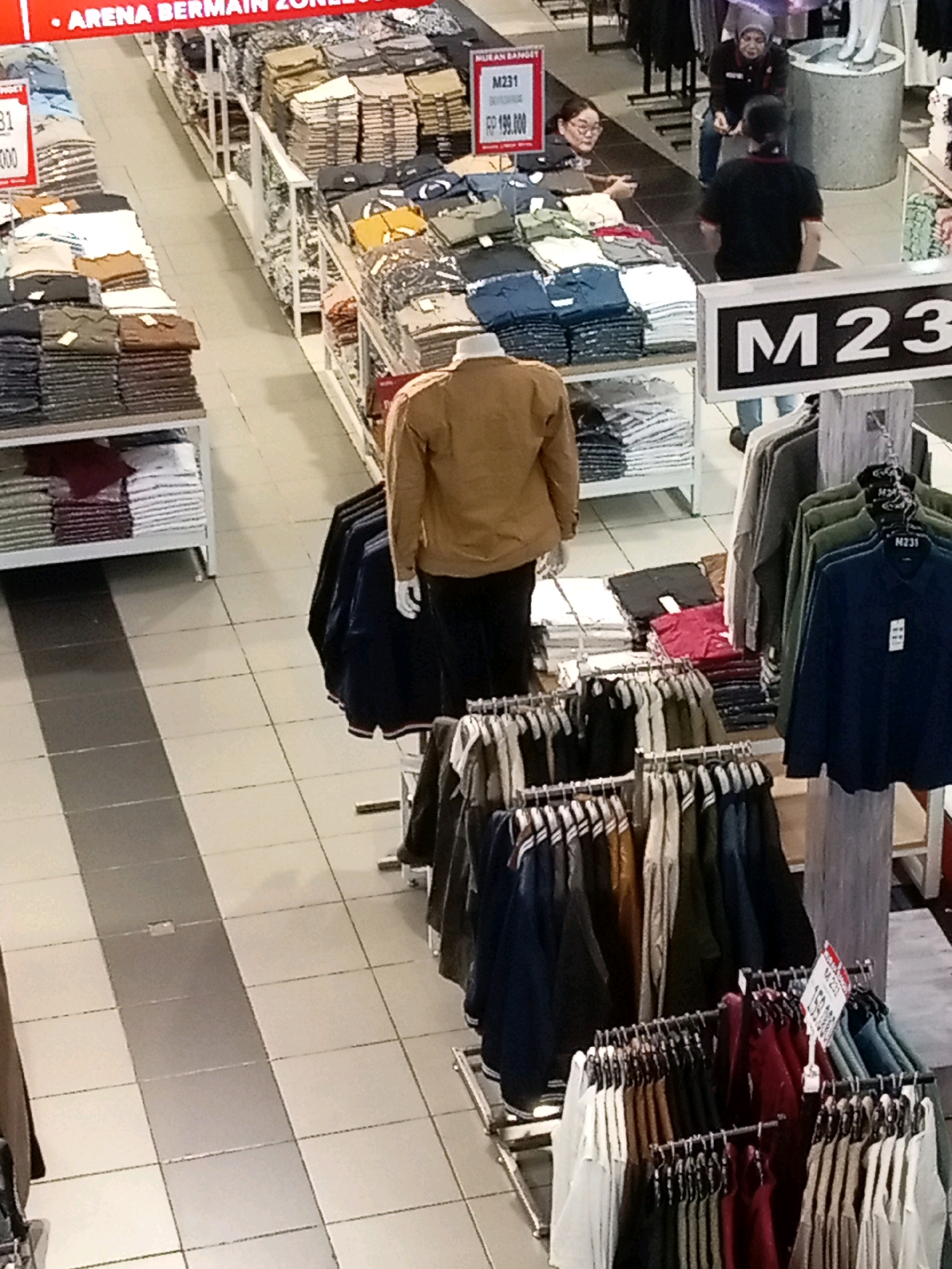 An aerial, high-angle shot shows the interior of a clothing store in Batam, Indonesia, with a focus on displays of folded and hung apparel. In the foreground, a mannequin wearing a tan jacket is positioned among racks of dark blue, green, and grey long-sleeved shirts. To the left, stacks of folded t-shirts and collared shirts in various colors, including white, brown, and dark grey, are arranged on shelves. A red and white price tag reading "M231" and "Rp 199.000" is visible on a display of folded shirts. In the midground, two women and one man are visible. One woman, wearing glasses and a grey hijab, stands behind a counter and appears to be interacting with a man in a black t-shirt and jeans. Another woman, wearing a grey hijab and a black jacket, is seated behind them. The background reveals more clothing racks and displays, with a large sign displaying "M23" prominently on a white background. The store's floor is tiled with a pattern of light and dark grey squares. The lighting appears to be artificial, typical of an indoor retail environment.