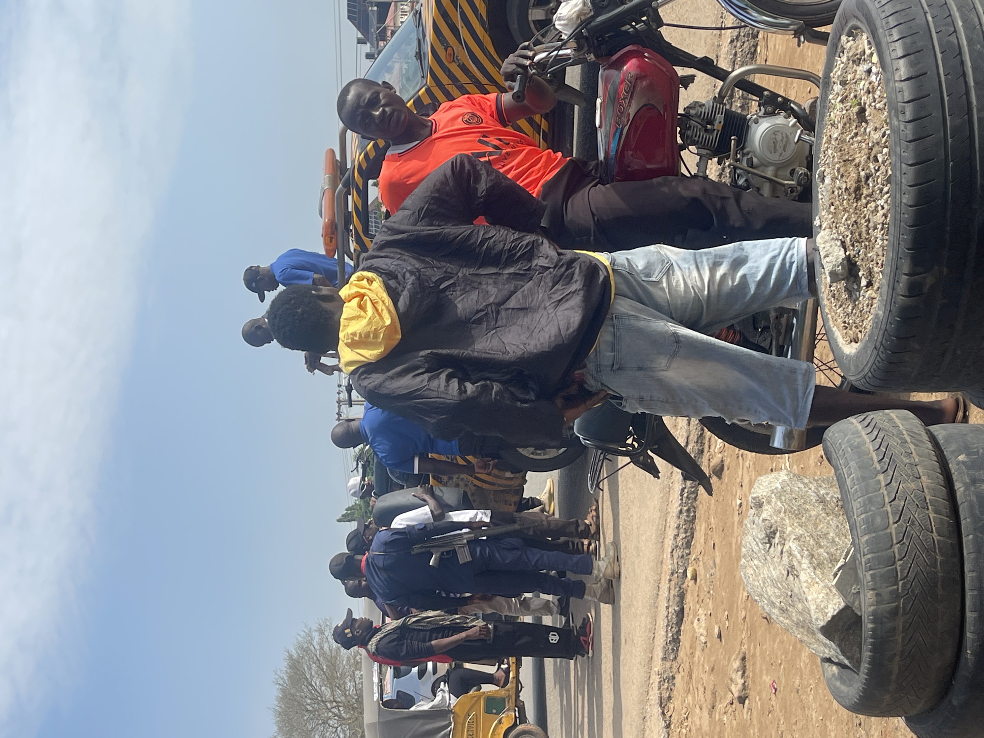 In Abuja, Nigeria, under a clear sky, a group of men are gathered near a roadside. The scene appears to be an informal gathering, possibly a roadblock or a waiting area, given the presence of a breakdown truck with a yellow and black hazard pattern. In the foreground, stacked tires serve as makeshift planters, holding soil and rocks.
Several individuals are the focus of the image. One man, wearing a black jacket over a yellow hoodie and light blue denim shorts, stands facing away from the camera, his back to the viewer. To his right, a man in an orange t-shirt with a prominent graphic is seated on a red motorcycle, looking towards the camera. His posture suggests he might be interacting with the person taking the photo or someone nearby.
Other men are dispersed around the scene. One man in a dark blue uniform with a rifle slung over his shoulder stands prominently, facing left. He is wearing dark pants and light-colored boots. Another man, in a red shirt and black pants, stands to his left, observing the surroundings. A yellow motorized tricycle is visible on the far left of the frame, with a person seated inside. In the background, several other individuals can be seen, some standing, some possibly on or near other vehicles. The ground is a mix of dirt and some debris, typical of a roadside environment in many parts of Abuja. The overall atmosphere is one of casual assembly with a potential underlying purpose that is not immediately clear from the image alone.