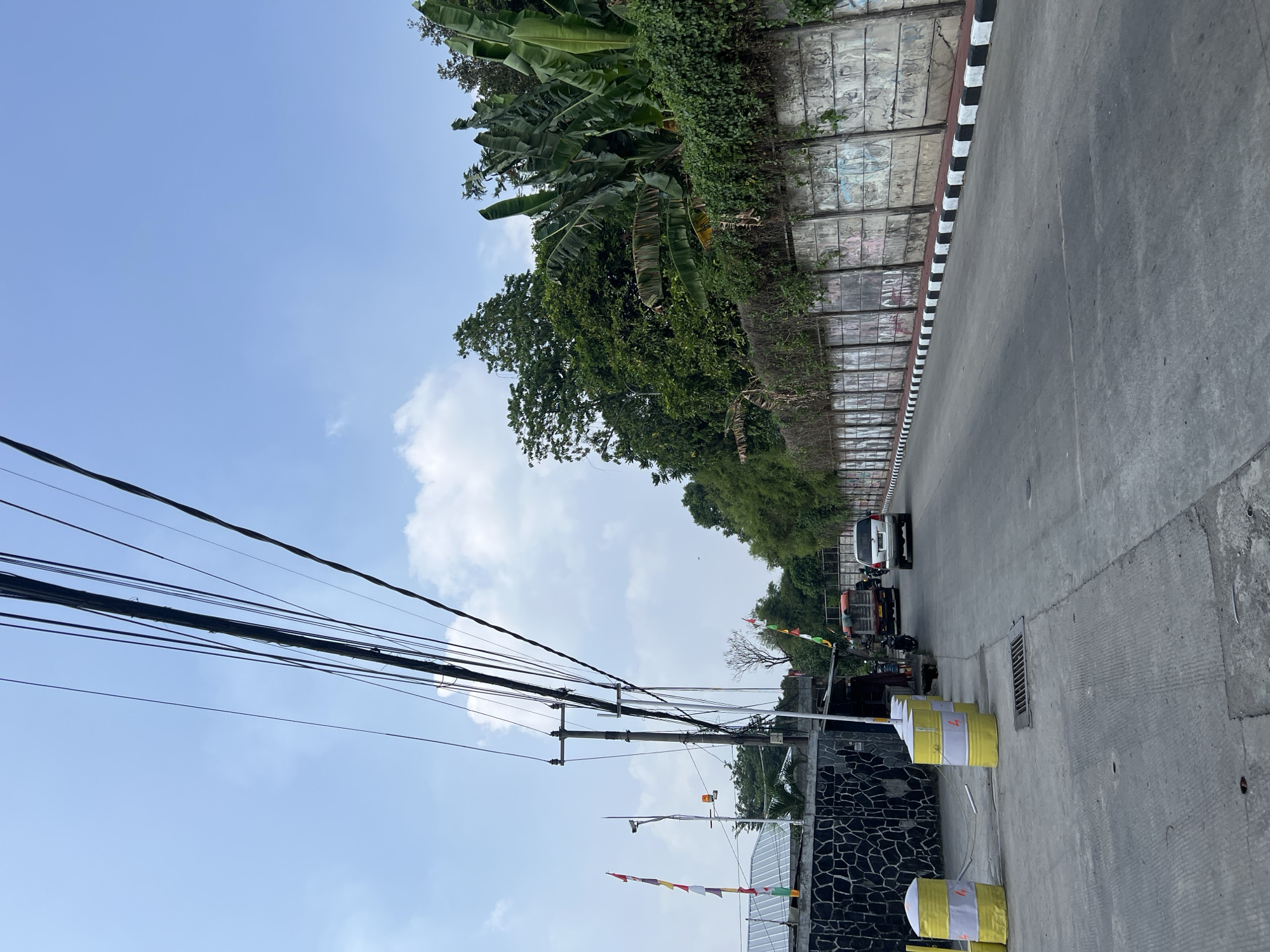This is a street-level, eye-level, medium shot of a paved road in Gunung Putri, Indonesia, under a clear blue sky with scattered clouds. The road runs diagonally from the bottom right to the top left of the frame. On the right side of the road, there is a concrete retaining wall covered in graffiti, with lush green vegetation, including banana trees, growing on top of it. The left side of the road features a dark stone wall, a small building with a corrugated metal roof, and several bright yellow cylindrical barriers with white stripes and lightning bolt markings. A utility pole with numerous black cables stretching across the sky is prominent on the left side. In the distance, a white SUV is driving away from the camera, followed by a red truck. Several small Indonesian flags are displayed on poles near the stone wall. The scene appears to be an urban or semi-urban environment with evidence of ongoing development or maintenance, as indicated by the construction barriers.