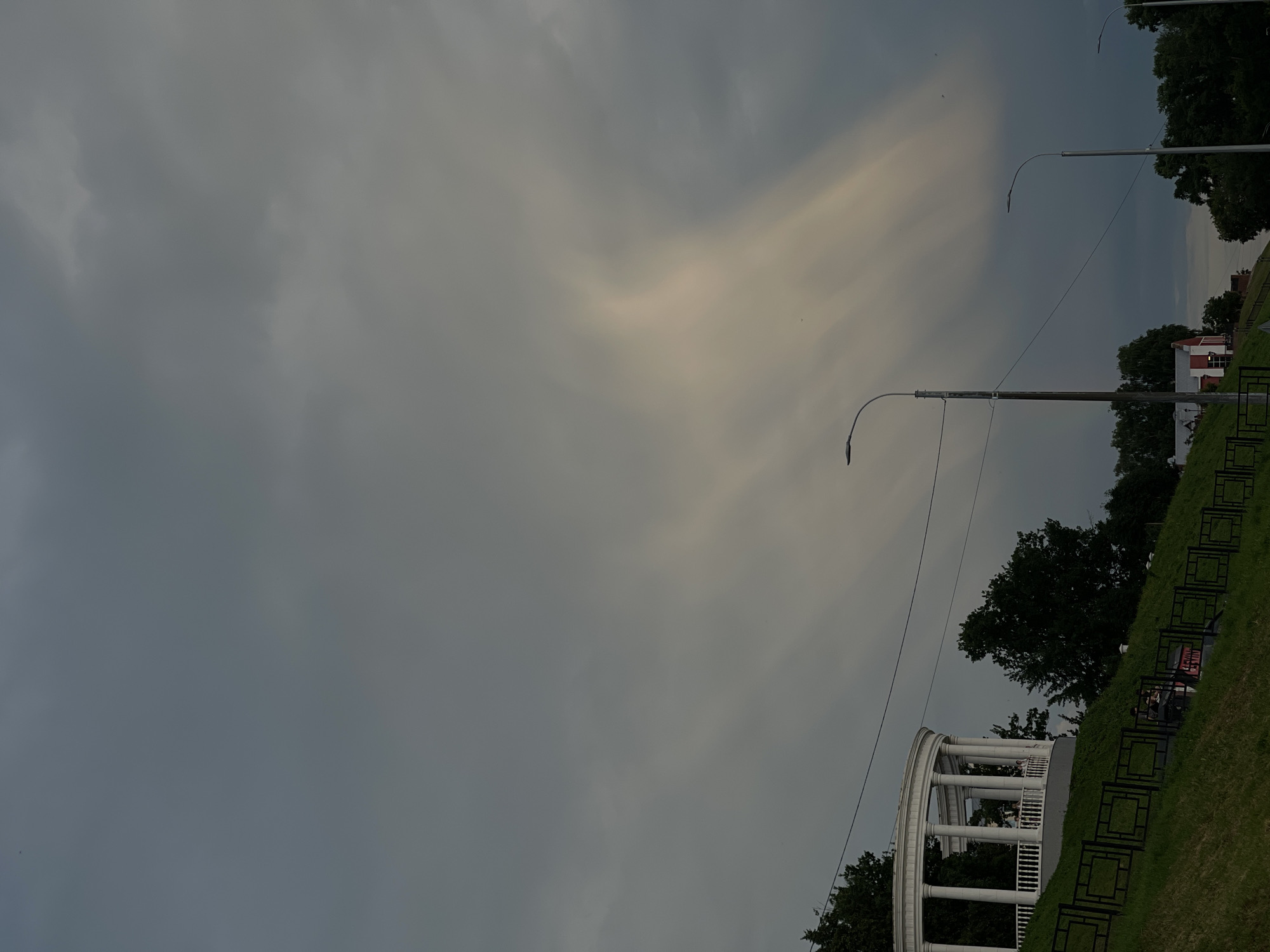 The image depicts a cloudy sky above a grassy embankment in an urban setting. On the left, a white, circular gazebo with columns and railings sits atop the embankment. A black metal fence runs along the edge of the embankment, with a few people visible near it. Several trees are scattered on the embankment and in the background. Three streetlights are present, with the one closest to the viewer being the tallest. In the background, a small red and white building is visible. The overall lighting suggests it is either dawn or dusk.