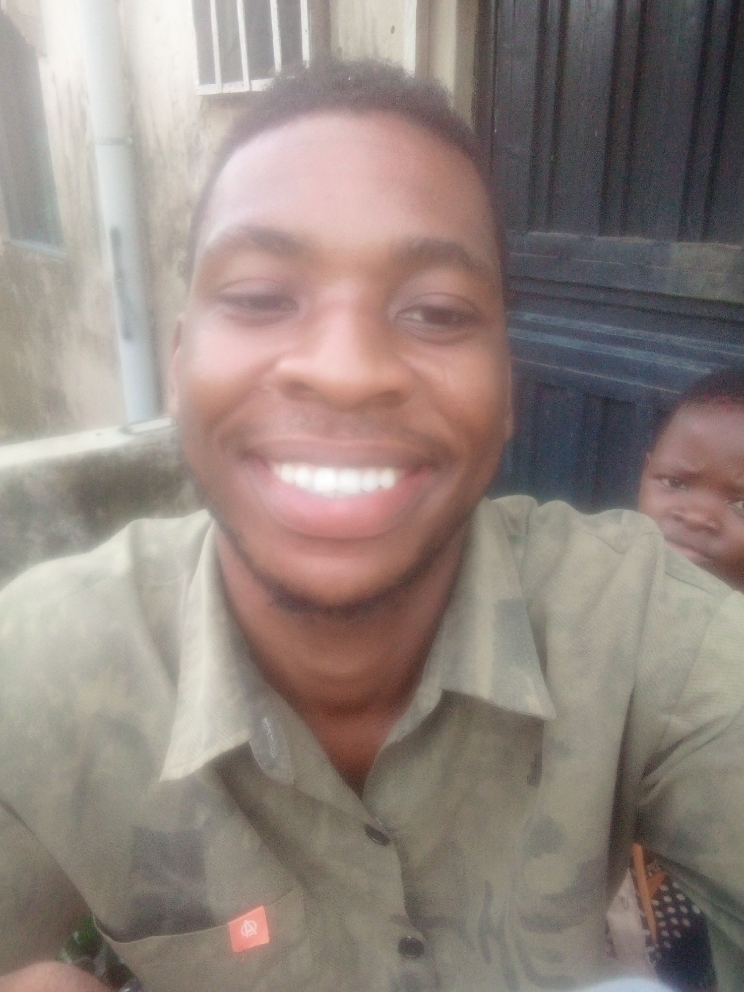 A selfie captures a man smiling broadly at the camera in the foreground, with a child partially visible behind his right shoulder, also looking towards the camera. The man has short, dark hair and is wearing a light olive green collared shirt. A small, orange rectangular tag with a white stylized 'A' or abstract logo is affixed to the left chest pocket of his shirt. His wide, open-mouthed smile reveals bright white teeth, suggesting a cheerful or happy emotion. The child behind him appears to have a neutral or slightly curious expression.
The scene is set outdoors or in a semi-enclosed exterior space, illuminated by natural, diffused daylight. In the background, to the left, parts of a light-colored building wall, a white vertical downspout, and a window with white bars are visible. To the right, a dark, vertically slatted surface, possibly a door or gate, forms another part of the backdrop. The image has a slightly soft focus, particularly noticeable in the background elements.