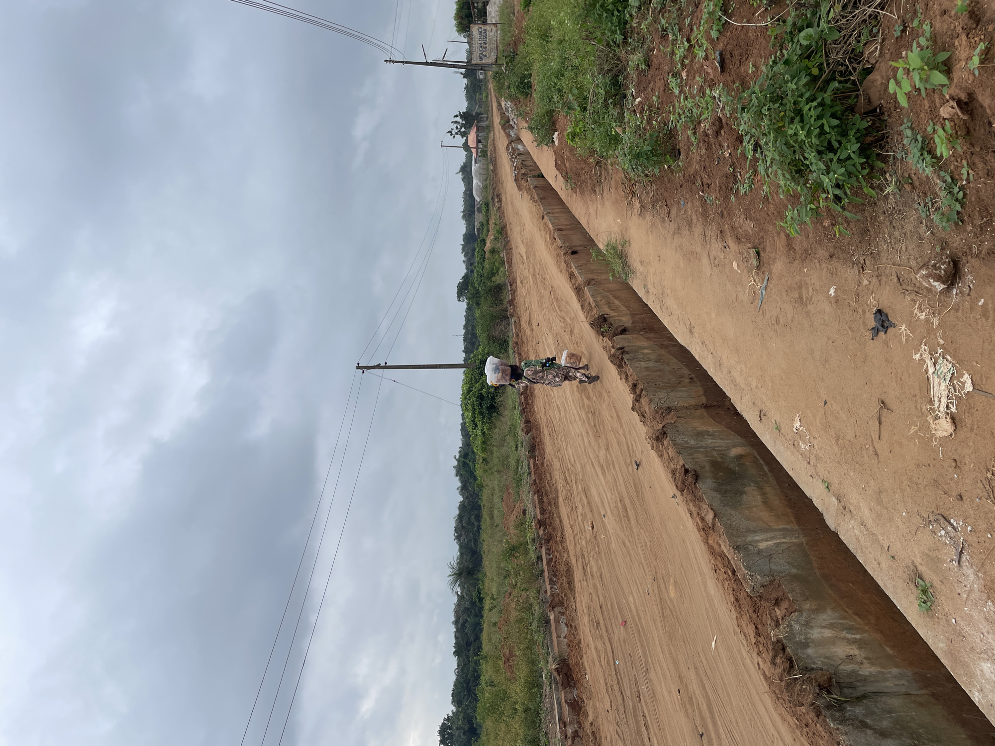 In Iyafin, Nigeria, a person walks down a dirt road carrying bundles on their head and in their hands. The road is flanked by a concrete drainage ditch on the left and overgrown vegetation on the right. Overhead, power lines stretch across a cloudy sky. In the distance, there are trees and structures, including a sign for the "ANGLICAN CHURCH OF THE MISSION." The scene depicts a rural or semi-rural setting with evidence of ongoing development or road maintenance.