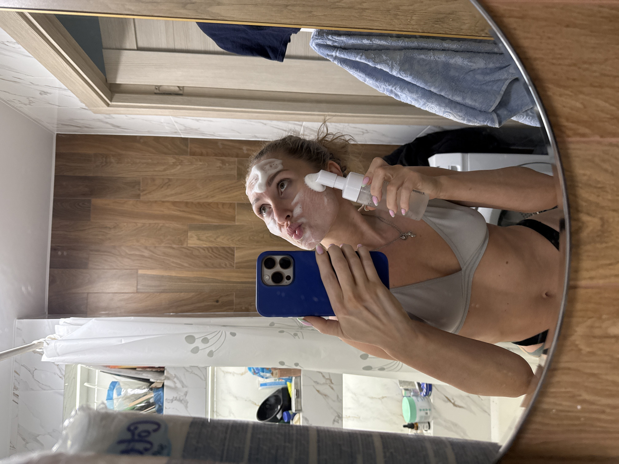 A woman with blonde hair pulled back is in a bathroom, reflected in a large mirror. She is wearing a grey sports bra and black underwear. Her face is covered in white foam, and she is holding a white bottle with a pump dispenser to her face, appearing to apply more foam. She is holding a blue smartphone in her left hand, taking a selfie. The bathroom has white marble-patterned tiles on the walls and wooden plank-style paneling. A white shower curtain with a floral pattern is visible. In the reflection, a washing machine, a blue towel, and various bathroom toiletries are present. There are no indications of the city of Perm, Russia.