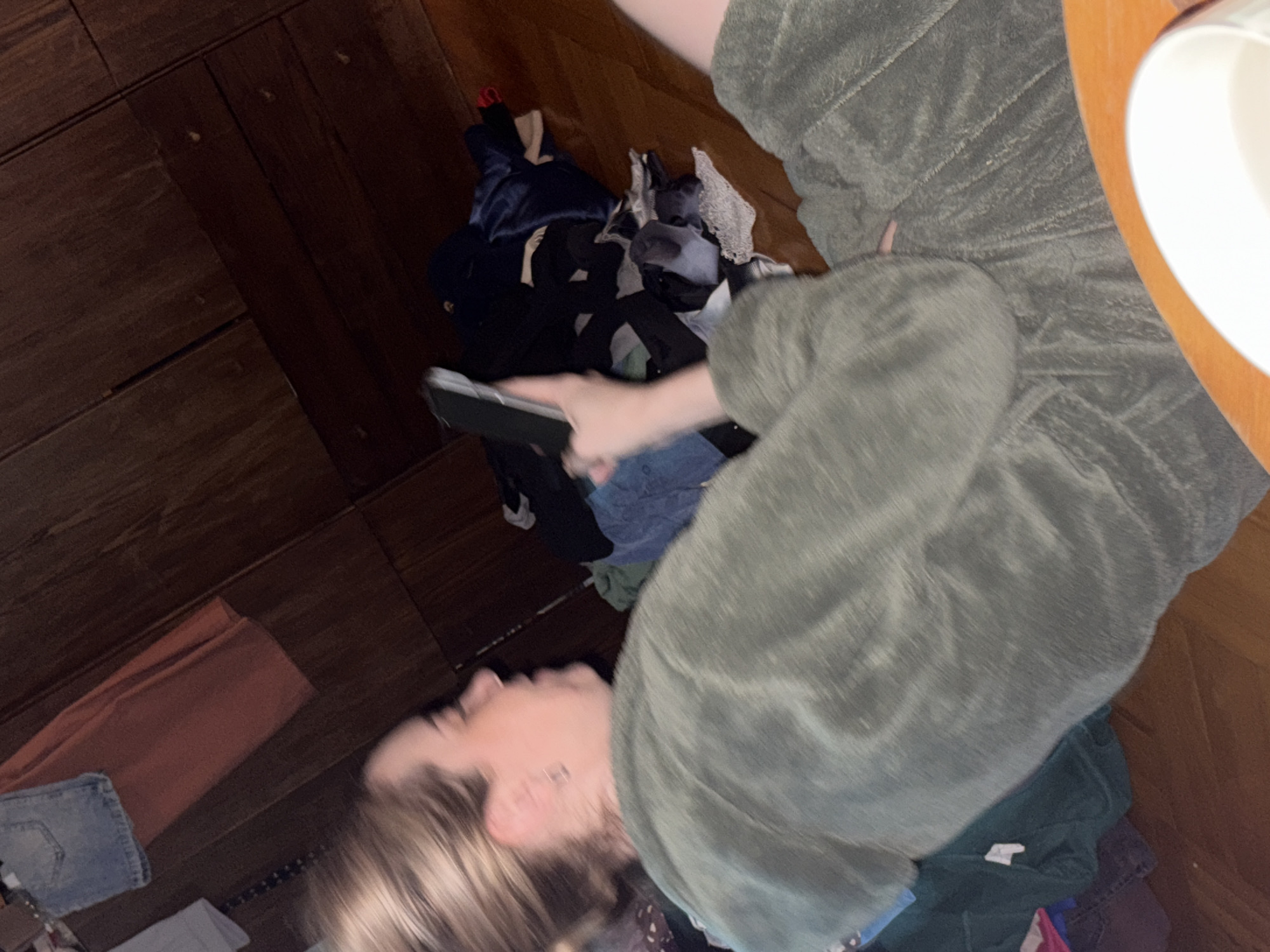 A woman is lying on a wooden floor, holding a smartphone in her hand, seemingly interacting with it. A pile of clothes is near her, and a small table with a white mug on it is partially visible in the upper right corner of the frame. The location is unspecified, and there is no information to support the image being in Sinyavino, Russia.