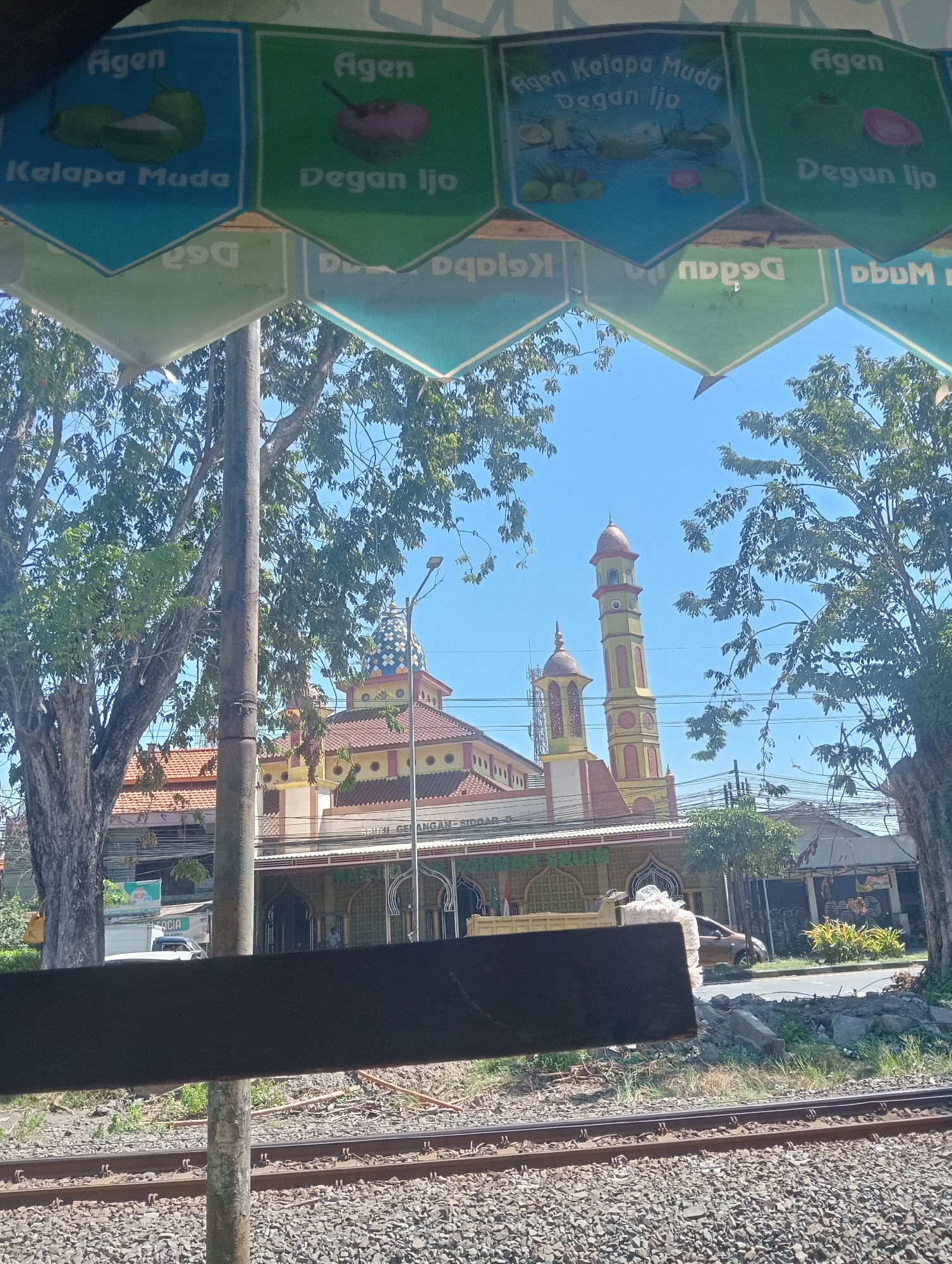 The image captures a view of a mosque in Sruni, Indonesia, framed by a canopy displaying advertisements for "Kelapa Muda" (young coconut) and "Degan Ijo" (green coconut). The mosque has a red-tiled roof and a distinctive yellow and green minaret topped with a patterned dome. In the foreground, railway tracks run parallel to a road where a truck and a car are visible. The sky is clear and blue, with lush green trees flanking the scene. The overall impression is of a typical day in a tropical Indonesian town.
