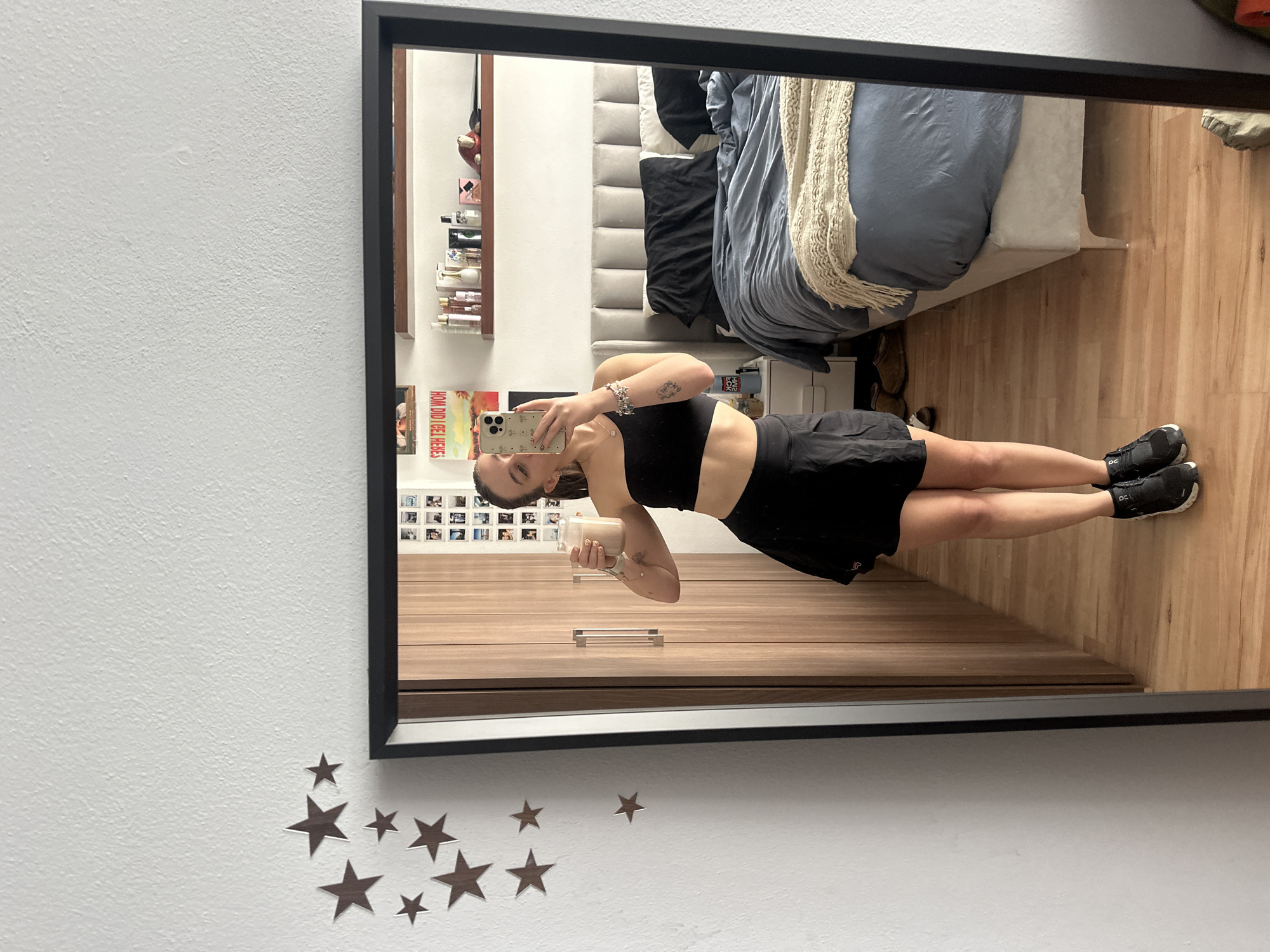 A woman in a black sports bra and black skirt is taking a selfie in a full-length mirror. She is holding a jar with a light-colored substance inside. The background includes a bed with a gray duvet and a beige throw blanket, a wooden floor, and a wooden cabinet. Above the cabinet are a shelf with bottles and other items, and a wall with framed photographs. The mirror is rectangular with a black frame and hangs on a white wall decorated with star-shaped stickers. The location appears to be indoors, possibly a bedroom. The city of Huixquilucan de Degollado, Mexico, cannot be confirmed from the image alone.
