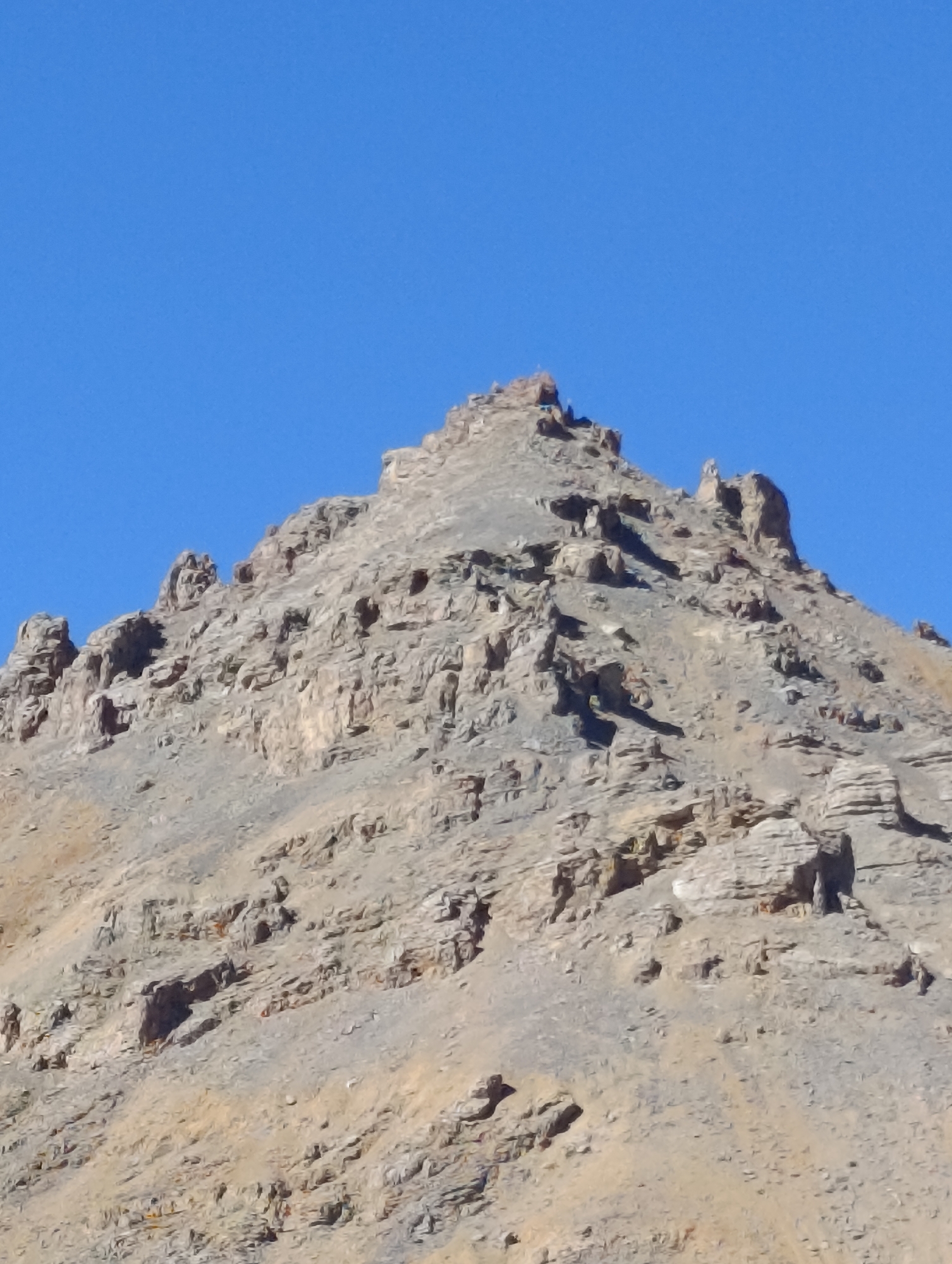 A close-up, upward-facing shot shows the jagged, rocky summit of a mountain under a clear, bright blue sky. The mountain's surface is rough and textured, with layers of stone and scree. Shadows are cast in various crevices, highlighting the uneven terrain. The peak is sharp and irregular, with several prominent rock formations jutting upwards. There are no visible signs of human presence or structures. The lighting suggests it is daytime, likely a sunny day.