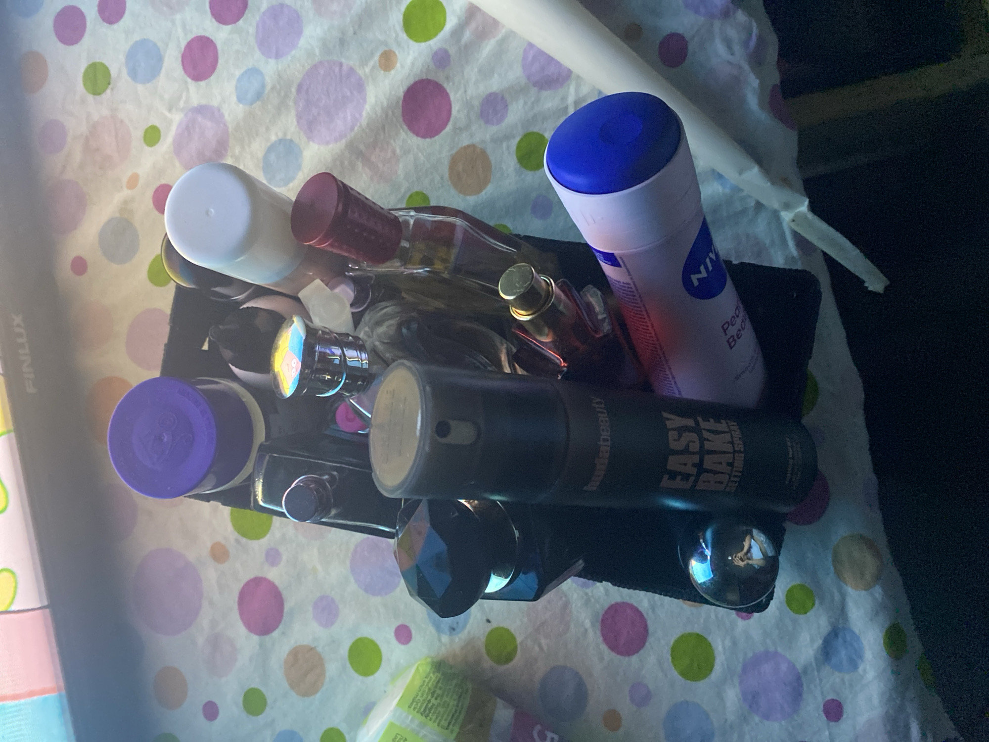 The image displays an overhead view of various cosmetic and beauty products arranged in a black organizer or tray. The items are set on a light-colored surface decorated with a pattern of colorful polka dots in shades of pink, green, blue, purple, and brown.
Key objects include several perfume bottles of different shapes and sizes, some with metallic or decorative caps. A prominent pink and white spray can with a blue cap is visible, featuring the brand name "NIVEA" and the text "Pearl & Beauty." Adjacent to it is a dark-colored spray can, clearly labeled "hudabeauty EASY BAKE SETTING SPRAY." Another product with a distinctive purple cap is situated in the left part of the organizer.
The lighting in the image creates reflections and glare on the glossy surfaces of the bottles and the polka-dotted background. A notable detail, observed in a cropped section, is the reflection of a person (likely the photographer) in a spherical, faceted object within the organizer, showing the upper torso and head. Partial text "FINLUX" is also visible on packaging on the far left. The setting appears to be indoors, potentially a personal vanity or bathroom counter. No specific activity is occurring; the items are presented in a stored arrangement.