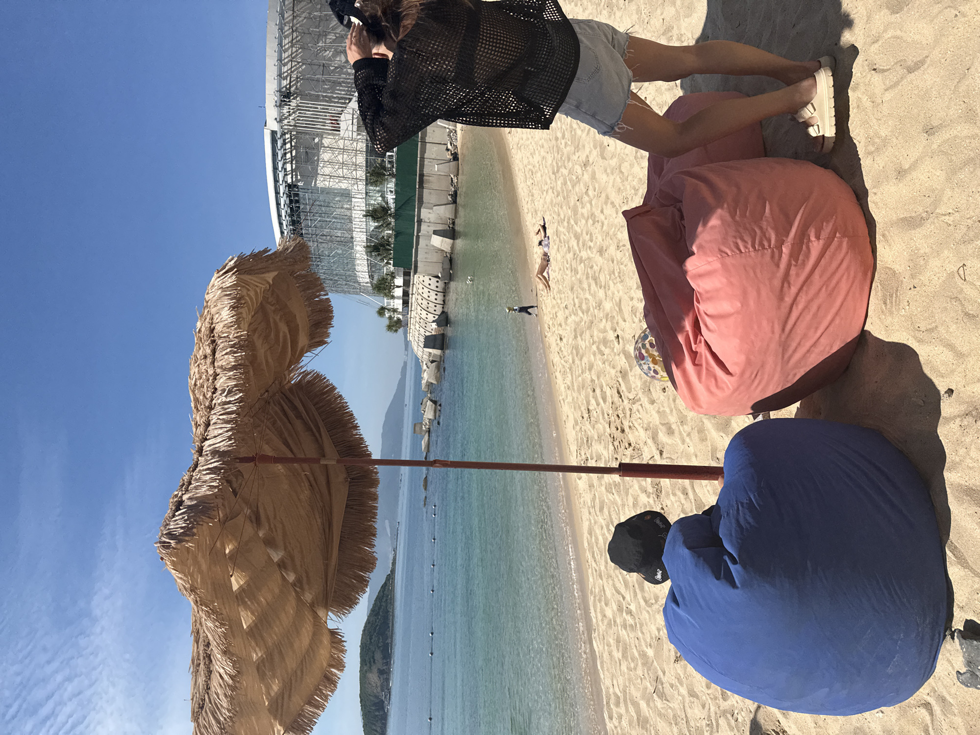 This image captures a scenic view of Nha Trang's beach in Vietnam. In the foreground, two large beanbag chairs, one blue and one pink, sit on the sand. A thatched umbrella shades the chairs, its fringe swaying gently. A person stands in the middle ground, facing away from the camera, wearing a black mesh top and denim shorts, and adjusting their hair. Further down the beach, two individuals are sunbathing. The turquoise water of the sea stretches out, dotted with a few buoys and leading to a distant island. In the background, a modern, partially constructed building with a glass facade rises above the coastline, contrasting with the natural beauty of the beach and sea. The sky is clear and blue, indicative of a sunny day.