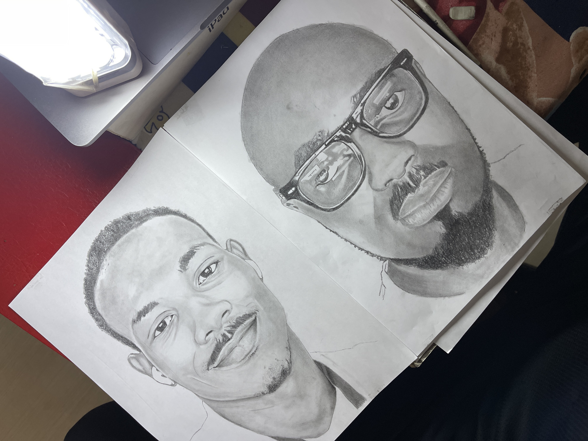 Two pencil portraits on white paper are positioned side-by-side on a table. The portrait on the right depicts a bald man with a beard, wearing glasses. The portrait on the left is of a younger man with short hair and a mustache. The drawings are detailed and shaded. The table surface is partially visible, showing red and dark surfaces. A laptop or tablet with a bright light source is in the upper left corner. Another object, possibly a cushion or pillow, appears in the upper right. The location in Jos, Nigeria is not inferable from the image.