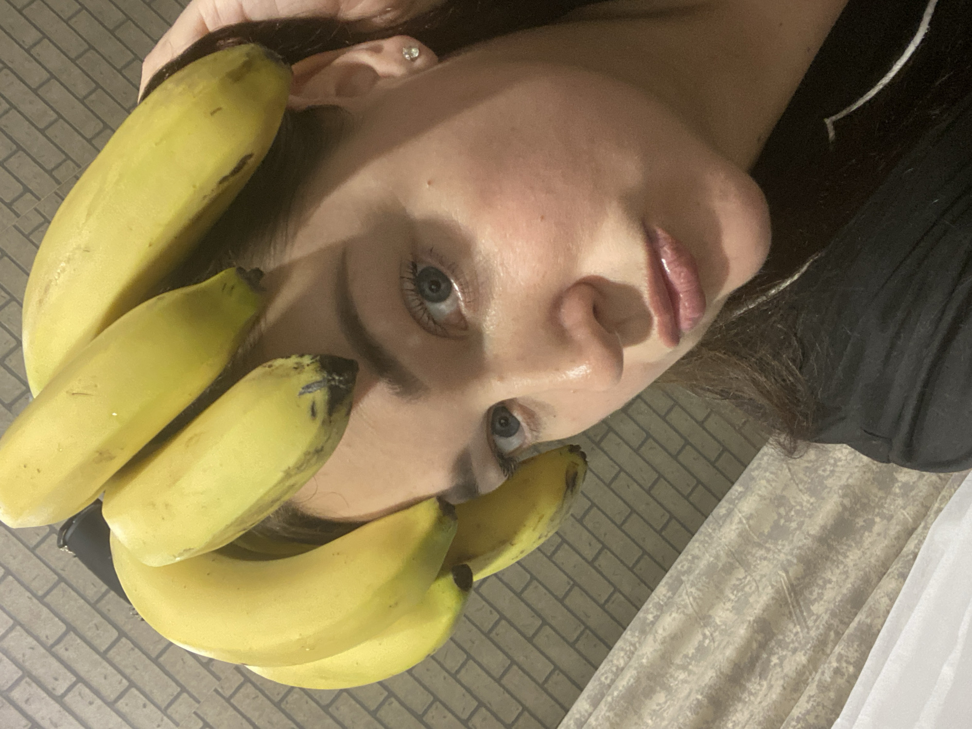 A close-up, high-angle portrait shows a woman lying on her back, looking directly at the camera. The image is rotated 90 degrees clockwise, positioning her head on the right side of the frame. A prominent bunch of ripe, yellow bananas, displaying some green hues and dark spots, is arranged around her head and face, with several individual bananas fanning out from behind and along her cheeks.
The woman has fair skin, blue or grey eyes, and dark hair, visible both around the bananas and extending to the left side of the frame. A small, sparkling stud earring is visible in her right ear. Her lips have a glossy finish, and her expression is neutral and direct, engaging the viewer.
The background consists of two main textures. To the upper left, a grey, rectangular brick-like pattern, likely a tiled floor, is visible. To the lower right, beneath her hair and shoulder, is a lighter, mottled fabric, possibly bedding or a curtain. She is wearing a dark, possibly black, garment. The soft lighting suggests an indoor setting.
The image presents a creative or artistic arrangement, playfully combining human features with natural elements. The location is Sochi, Russia. No text is visible in the media.