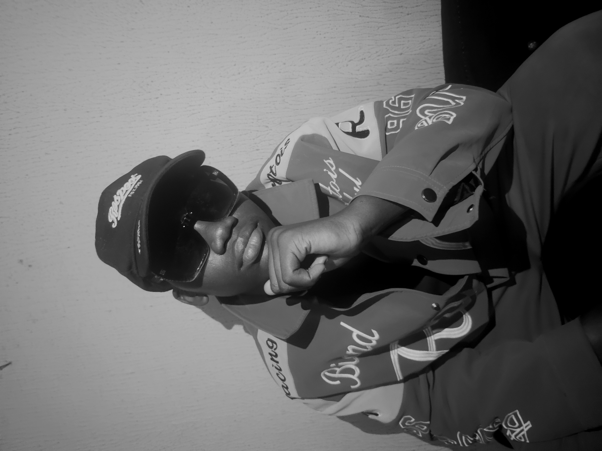 A black and white, vertically oriented image captures a person in a three-quarter profile, looking towards the right. The individual, appearing to be male, wears a dark baseball cap with the word "Respect" visible on the front, along with dark sunglasses that obscure their eyes. Their right hand is raised, partially covering their mouth and chin area.
The person is dressed in a long-sleeved jacket featuring various textual and graphic designs. Visible text elements on the jacket include "acing" (likely part of "Racing"), "Bind" in a cursive font, and "tois" (possibly part of a longer word). Stylized numbers or letters, such as "96," are also discernible. The jacket sleeves have buttoned cuffs. On the right side of the frame, part of the person's lower body, clothed in dark, plain trousers, is visible.
The background primarily consists of a plain, lightly textured wall on the left, suggesting an indoor setting. To the far right, a dark, indistinct object, possibly furniture, is present. Shadows are prominent, cast by the cap and head, indicating a distinct light source. The individual's pose and obscured face do not convey a specific emotion or activity beyond a staged portrait.