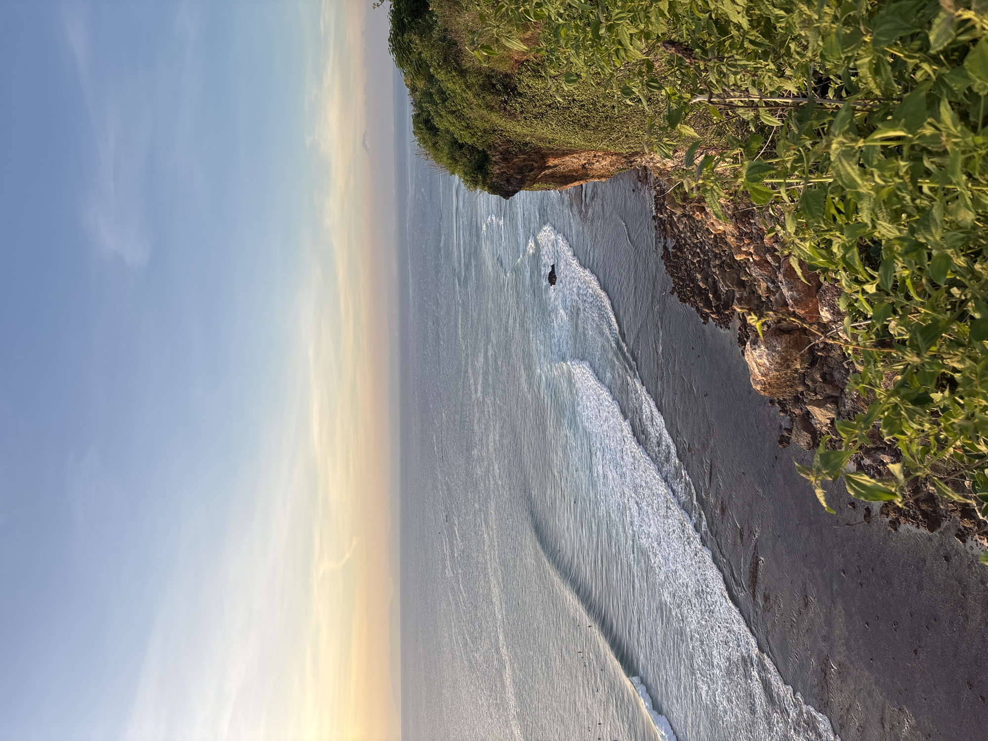 The image shows a coastal scene in Kuta Selatan, Indonesia. The view is from atop a cliff covered in green vegetation, looking out over the ocean. Waves are breaking along the shoreline, and the water appears to be a gray-blue color. In the distance, the sky is a mix of blue and pale yellow, suggesting either sunrise or sunset. A small rock formation can be seen in the water near the base of the cliff.