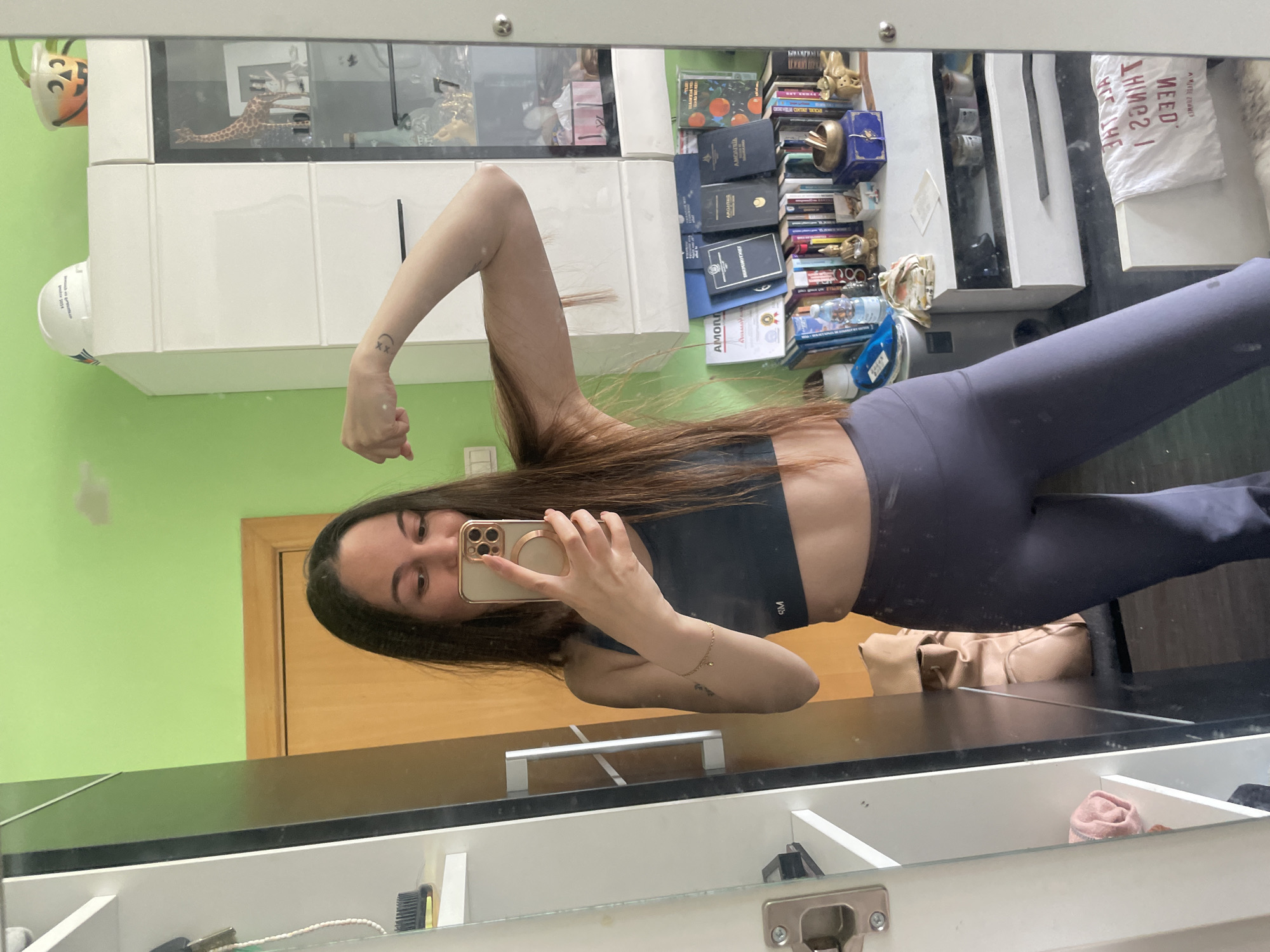 A female in athletic wear takes a selfie in a mirror. She is wearing a dark sports bra and dark leggings. The background includes a green wall, a wooden door, white cabinets, and a shelf with various items including books and a giraffe figurine. The location is not discernible from the image.