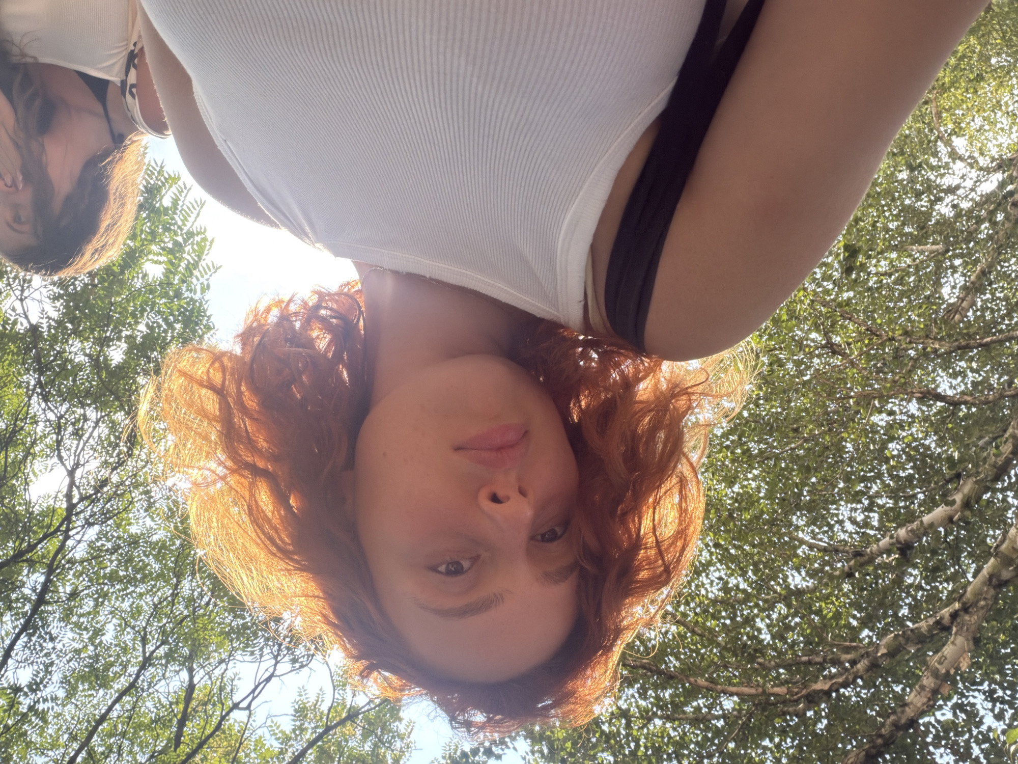 The image is a low-angle shot of a young woman with vibrant red, curly hair, illuminated by sunlight filtering through tree leaves. She is wearing a white ribbed tank top and a black strap is visible over her left shoulder. Behind her, another young woman with long brown hair is partially visible, covering her mouth with her hand. They appear to be outdoors, possibly in a park or wooded area, given the abundance of green foliage and tree branches overhead, suggesting a location in Illkirch-Graffenstaden, France, which is known for its green spaces. The sky is visible through the canopy of leaves. The overall lighting suggests it is daytime, with the sun positioned behind the main subject, creating a halo effect around her hair.
