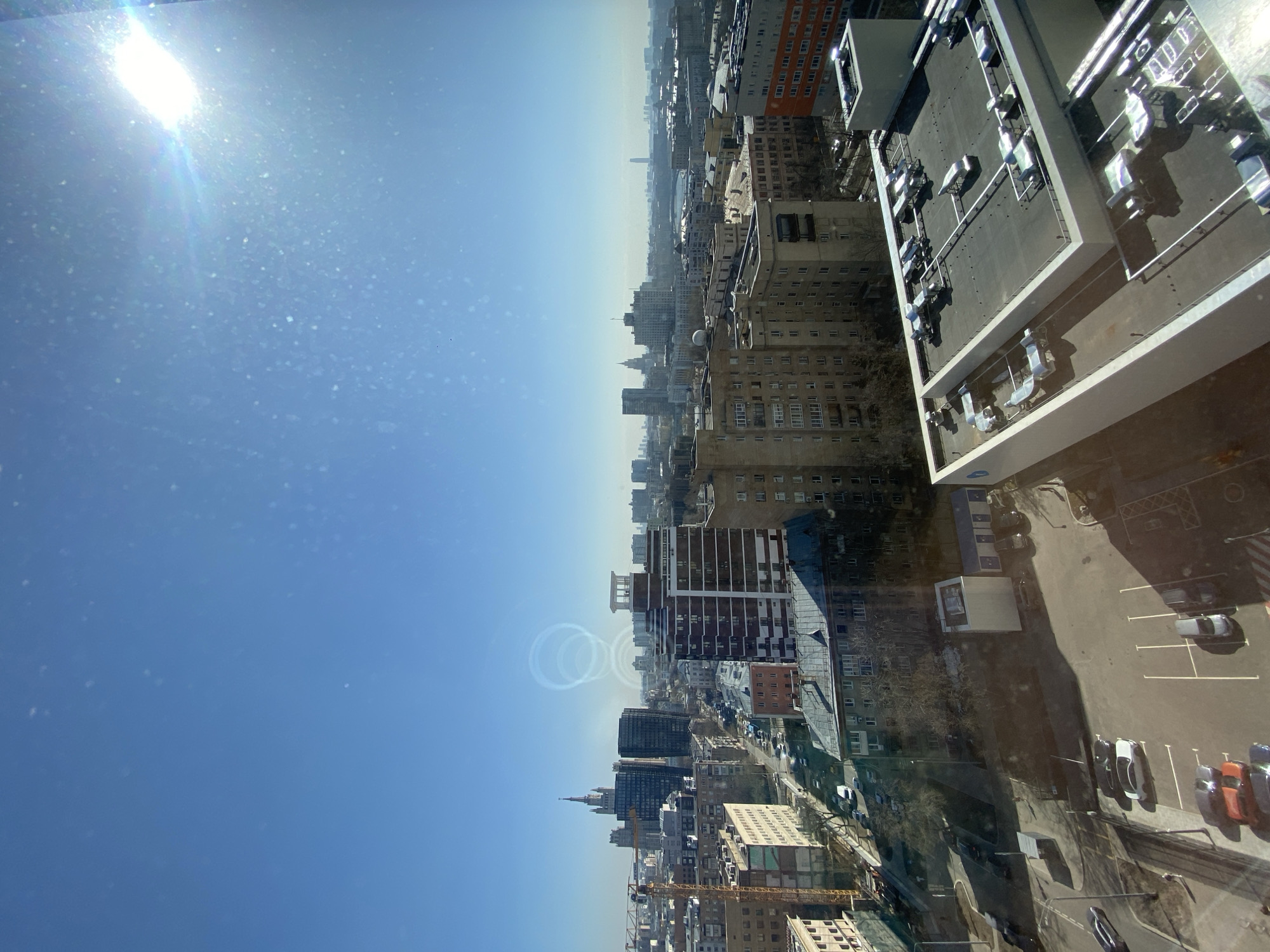 The image shows an aerial view of a city, identified as Moscow, Russia, on a sunny day. The top left of the image displays a clear, blue sky with the sun shining brightly. Below the sky, a dense urban landscape unfolds, including residential buildings, commercial structures, and infrastructure. The building on the right side is adjacent to a parking lot with several vehicles parked. A construction crane is visible among the buildings. The architectural styles of the buildings vary, with some featuring modern designs and others showing traditional Russian architectural elements.