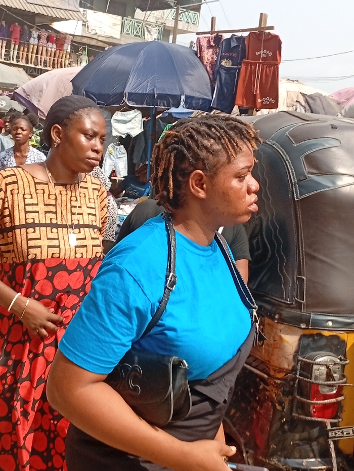 Two women stand in a bustling outdoor market. The woman on the left wears a patterned brown and red dress, a black head covering, and a delicate gold bracelet. The woman on the right sports a bright blue shirt over a black garment, with a black purse slung across her body. Both women gaze off to the right. Behind them, market stalls display clothing under blue umbrellas. A motorized tricycle is partially visible on the right. The scene is filled with activity and merchandise.