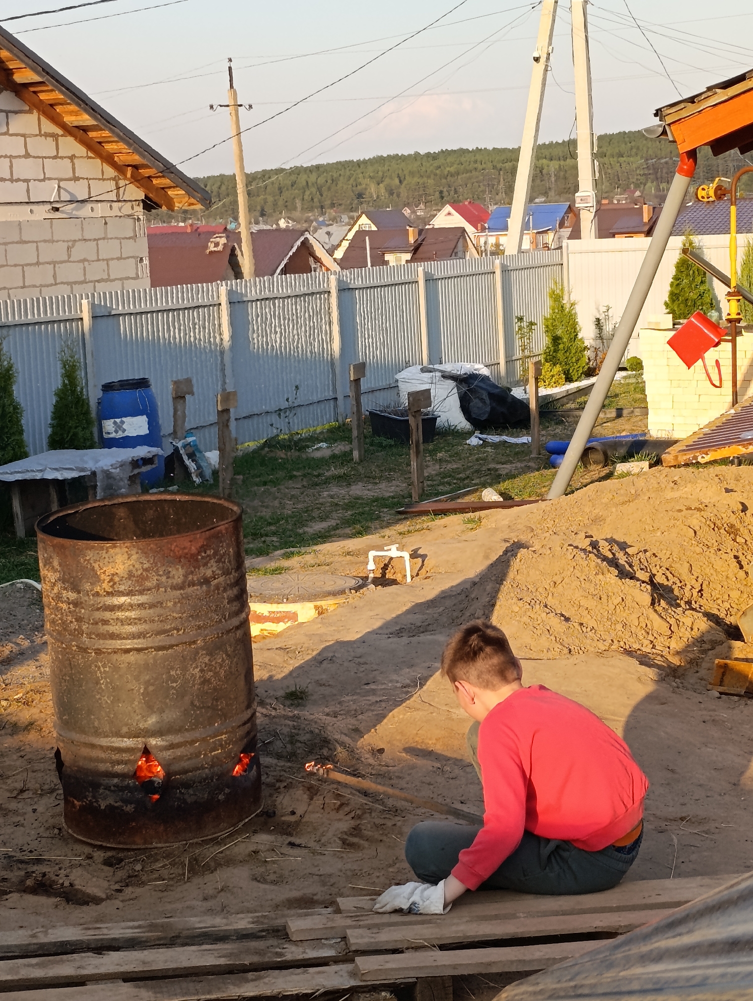 A young person sits on a wooden plank near a burning barrel at a site in the city of Chaikovsky, Russia. The barrel, which is rusty and has holes cut out of its lower section, contains a small fire. A mound of sand is next to the seated person. A blue barrel and a table are behind the barrel, near a corrugated metal fence. In the background are houses with various colored roofs and trees. An electrical pole is also visible.