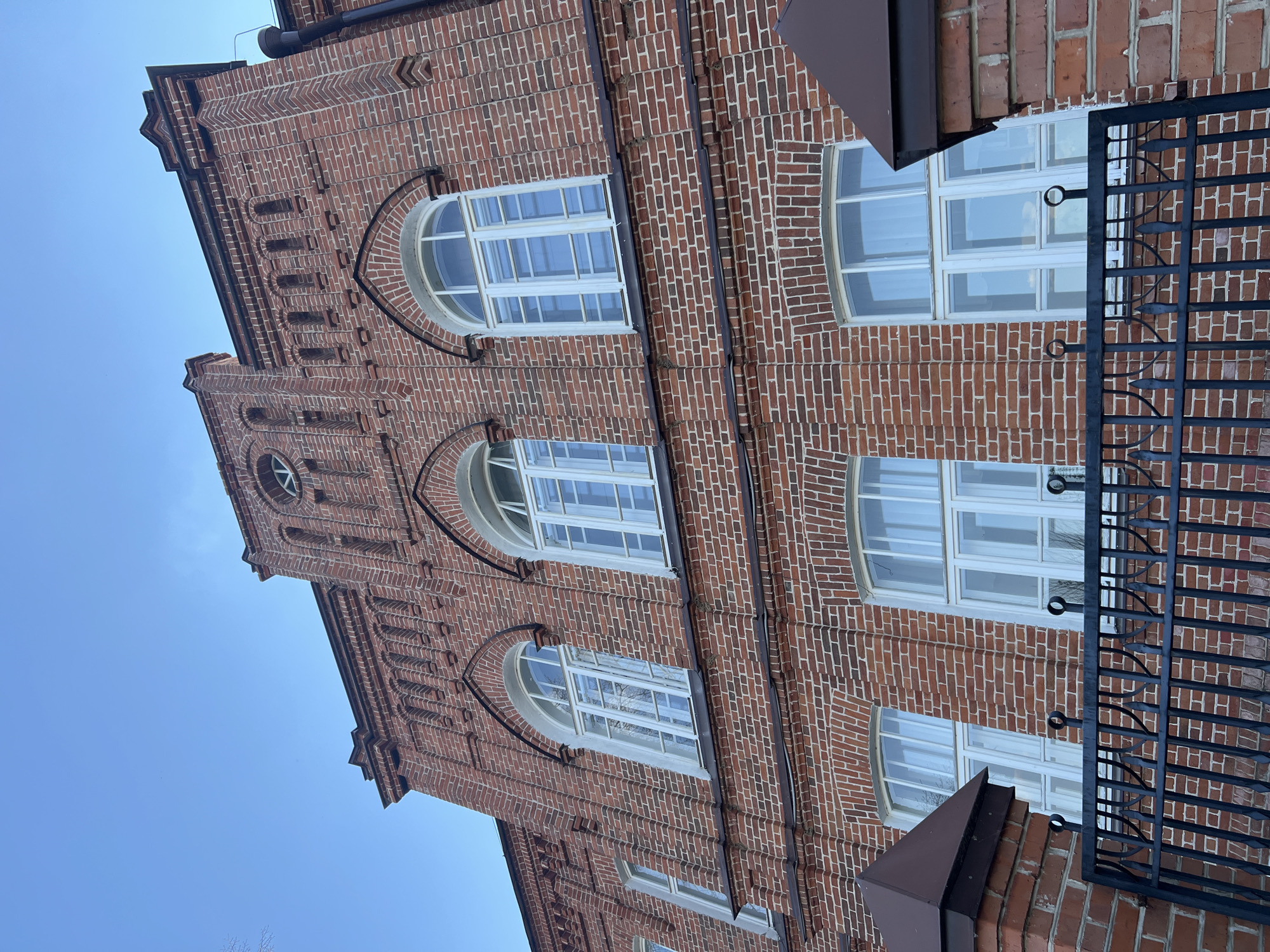 The image shows a section of the brick facade of a building in Sviyazhsk, Russia. The building is constructed of red brick and features arched windows with white frames. A black wrought iron fence is visible in the foreground on the right side of the image, partially obscuring the building's facade. The sky is a clear blue. The angle of the photo is looking upward, giving the building a towering appearance.