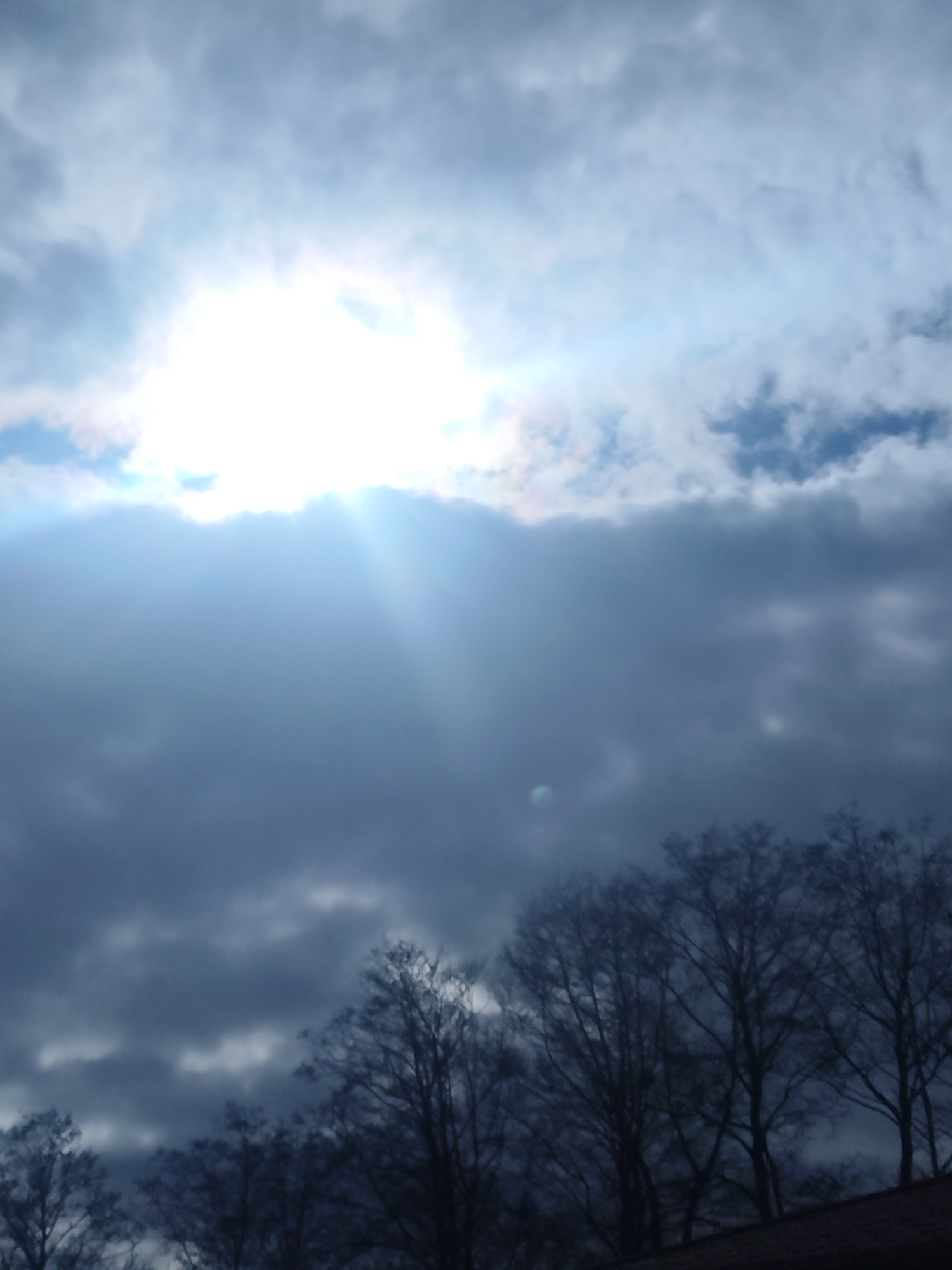 The image shows a view of the sky in what is likely Chaikovsky, Russia. The sky is filled with dark, gray clouds, with a bright patch where the sun is shining through. Rays of sunlight are visible through the clouds. Silhouetted trees are visible along the bottom of the image, suggesting a view from a residential or park area. The overall scene conveys a sense of overcast or stormy weather conditions.