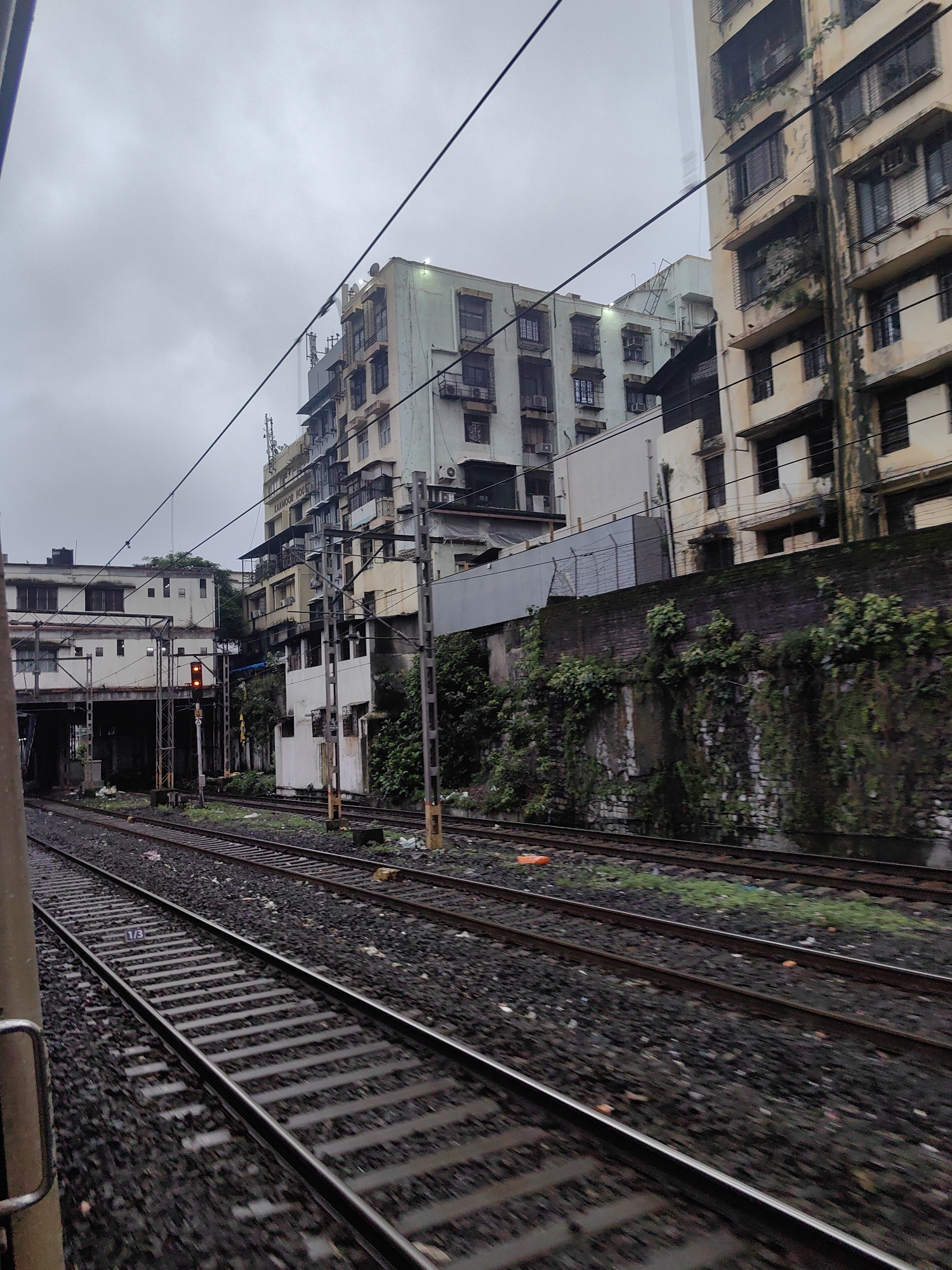 This is a medium-shot, exterior, ground-level, eye-level, overcast day photograph captured from a moving train, looking towards buildings and railway tracks in Mumbai, India. The scene depicts an urban environment with a focus on the infrastructure of a railway line.
In the foreground, multiple sets of railway tracks stretch out, with dark ballast and sleepers visible. The tracks are angled towards the left of the frame, suggesting movement or a curving line. The blurred motion of the train is evident in the streaks and soft focus of the foreground elements. To the far left, a portion of the train's exterior is visible, including a yellow pole and a silver handrail.
Behind the tracks, a weathered brick wall covered in moss and vegetation runs horizontally across the mid-ground. Above this wall, several multi-story buildings rise. The buildings are of varying designs and colors, with one prominently featuring a light green facade and numerous windows. Another taller building to the right appears older and more weathered, with a yellowish facade and visible balconies. The architecture suggests a densely populated urban area.
In the background, the sky is overcast and grey, contributing to the subdued lighting of the scene. A red railway signal is visible mounted on a pole near the tracks, indicating a stop or caution. Overhead, a complex network of electrical wires and poles crisscrosses the sky, essential for the functioning of the electric trains.
The environment appears to be a busy railway corridor within Mumbai, likely a suburban or main line, characterized by its dense development and active transportation infrastructure. No people are clearly visible in the frame, but the presence of the train and the buildings implies human activity and habitation. The overall atmosphere is one of a typical urban transit scene in India, characterized by its mix of modern and older infrastructure and the ever-present railway network.
