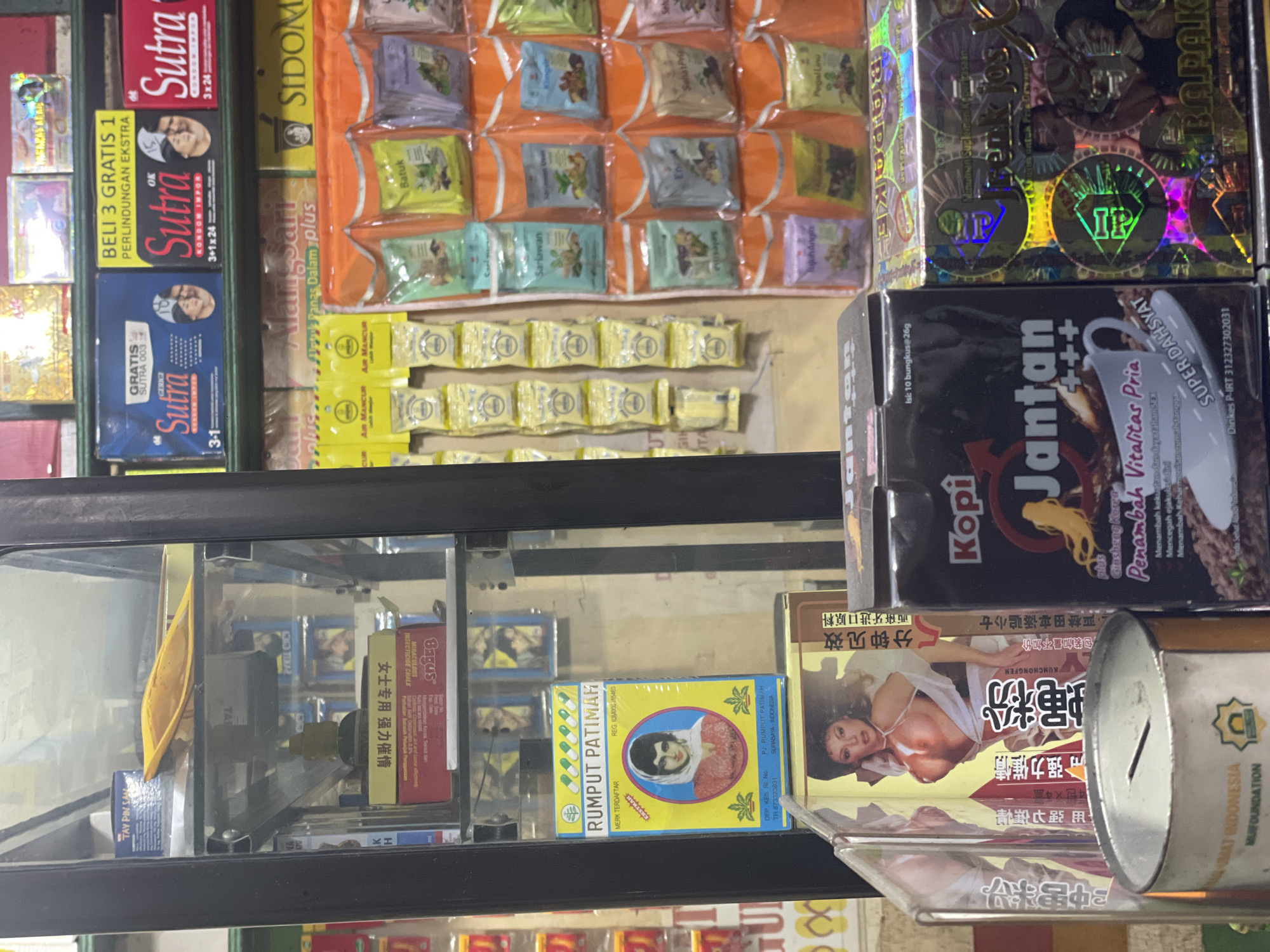The image shows a shop display in North Jakarta, Indonesia, filled with various products. At the top, there are rows of herbal remedies in small packets, along with boxes of "Sutra" condoms and a product called "Jantan Kopi" intended to enhance vitality. Below, within a glass display case, are several items, including a box of "Rumput Patimah" herbal medicine, a product with an image of a woman in suggestive attire, and a donation box with the logo of "Lembaga Amil Zakat Infaq dan Shodaqoh Nahdlatul Ulama" (LAZISNU). The display suggests a focus on traditional remedies, health supplements, and adult products.