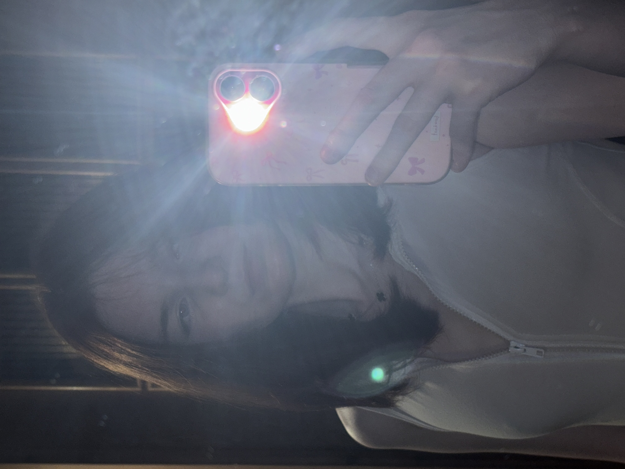 The image depicts a person holding a smartphone with a pink case adorned with bows, seemingly taking a selfie in a mirror. The flash is activated, causing significant glare. The person has dark hair and appears to be wearing a white top with a zipper. The environment is dark, potentially indoors, and reflections are visible on a surface, possibly indicating a window. There is no information available in the image that indicates the location is in the city of Vsevolozhsk, Russia.