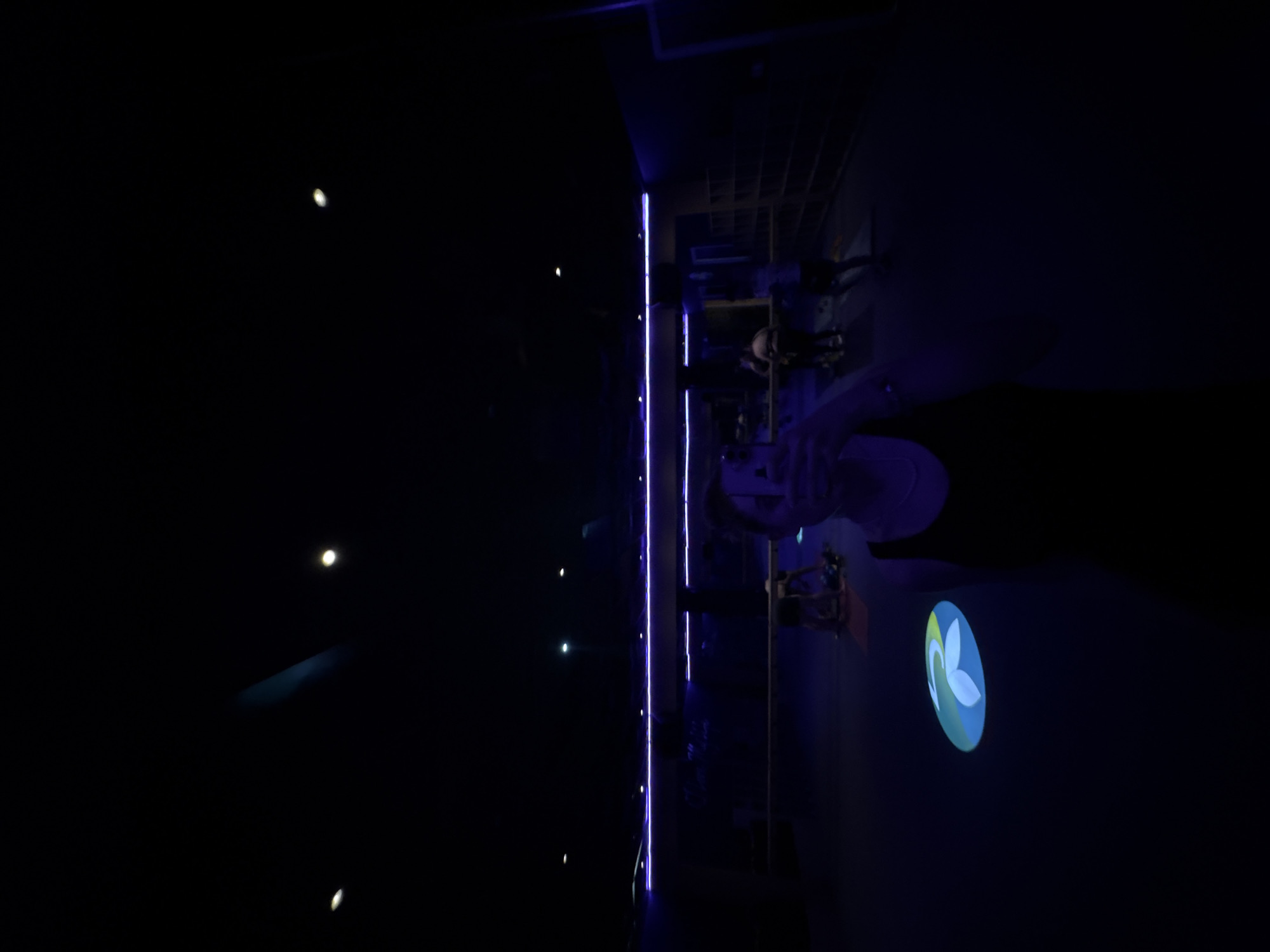 The image shows a dimly lit interior space in Mexico City, Mexico, with a distinct purple-blue lighting scheme. A vertical row of lights runs along a wall, casting a linear glow. In the darker area, there are a few small, scattered points of light. A logo, featuring a stylized floral or leaf design, is projected onto a wall. Figures of people can be distinguished, although their details are obscured by the darkness.