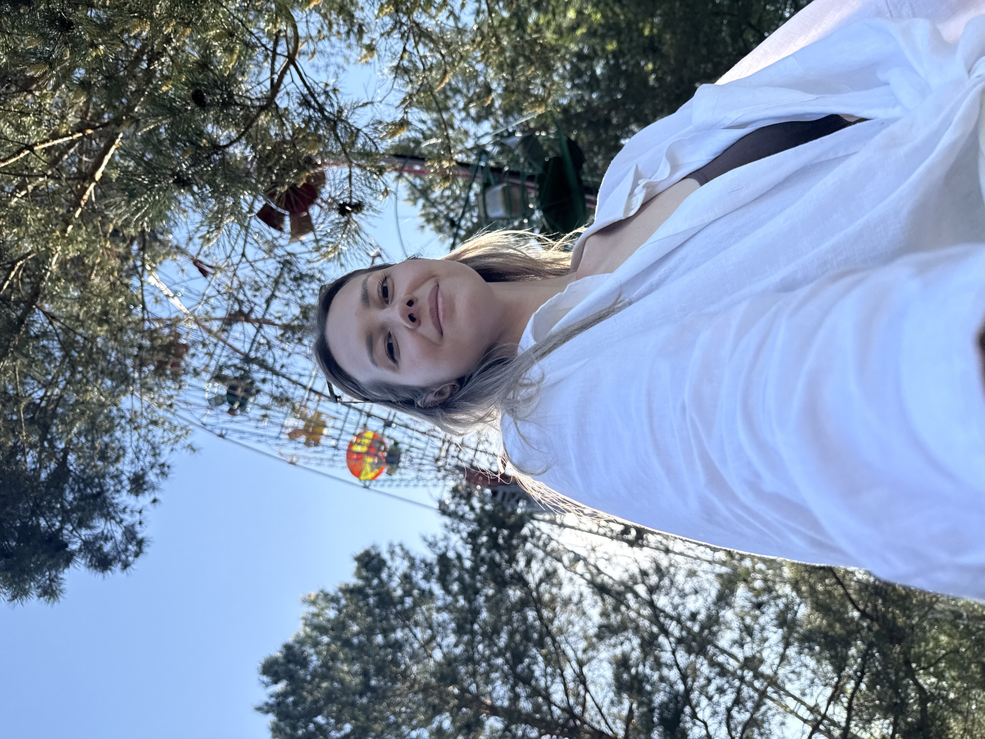 A low-angle shot shows a woman wrapped in a white garment looking down at the camera. The Ferris wheel is visible between the trees. The sky is blue. The location is Chaikovsky, Russia.