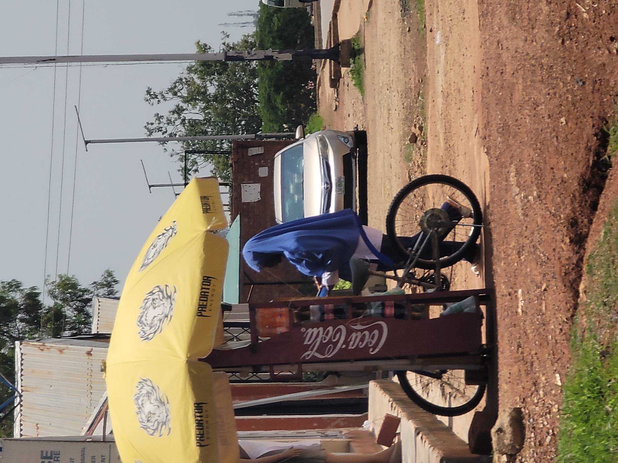 In Barakin Lerawi, Nigeria, a person wearing a blue hijab and white clothing stands beside a red Coca-Cola display rack attached to a bicycle. The display rack holds several bottles of beverages. A yellow umbrella with "Predator Energy" branding is open and shades the display and bicycle. In the background, a light-colored sedan is parked on a dirt road, with lush green trees and bushes behind it. A utility pole with black and white markings is visible to the right. The ground is covered in reddish-brown dirt and gravel, with some patches of green grass. The scene appears to be an outdoor roadside stall.