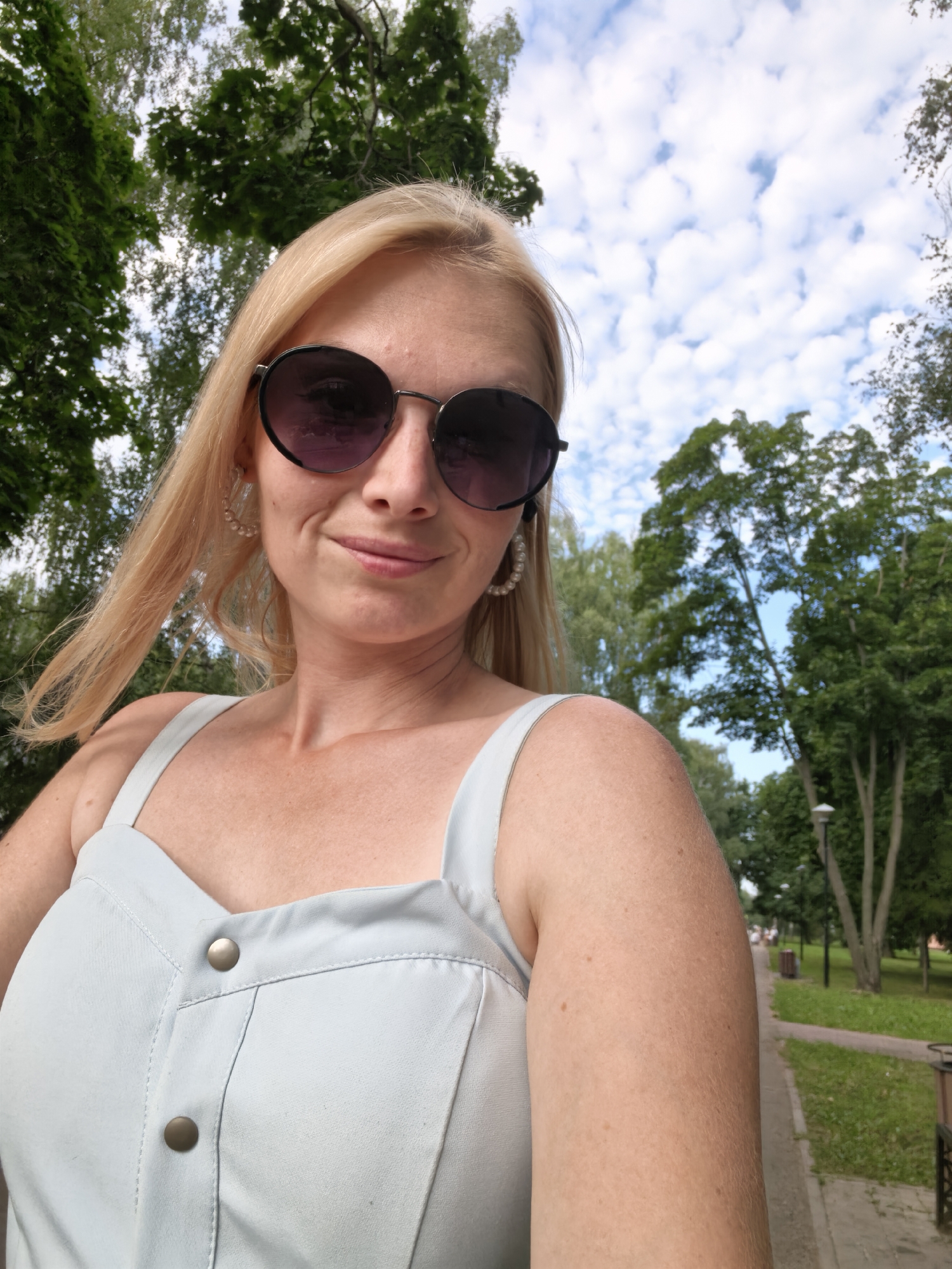 A woman with blonde hair and sunglasses takes a selfie outdoors. She is wearing a light blue dress with two buttons on the front. The background shows trees, a cloudy sky, and a park path. The woman appears to be smiling. The setting is likely Sterlitamak, Russia. The image suggests a daytime scene with pleasant weather.
