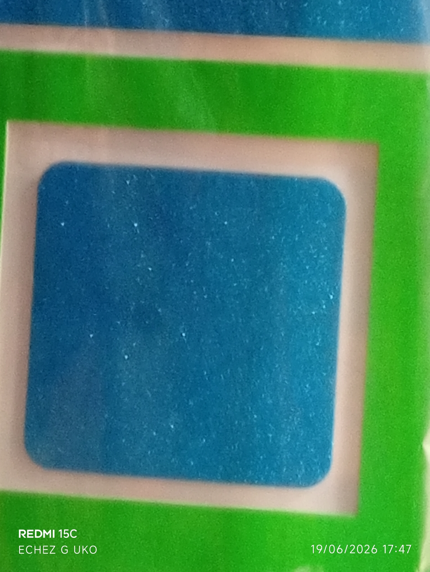The image is a close-up, slightly blurry photograph of a product packaging. The primary focus is a rectangular, bright blue object with rounded corners, which appears to be a patch or pad. This blue object is set against a cream-colored background that is bordered by a vibrant green stripe. At the bottom left of the image, text reads "REDMI 15C ECHEZ G UKO". At the bottom right, a date and time stamp reads "19/06/2026 17:47". The overall impression is of a product shot, possibly for retail or marketing purposes. The setting is not identifiable as Jalingo, Nigeria, or any specific urban environment.