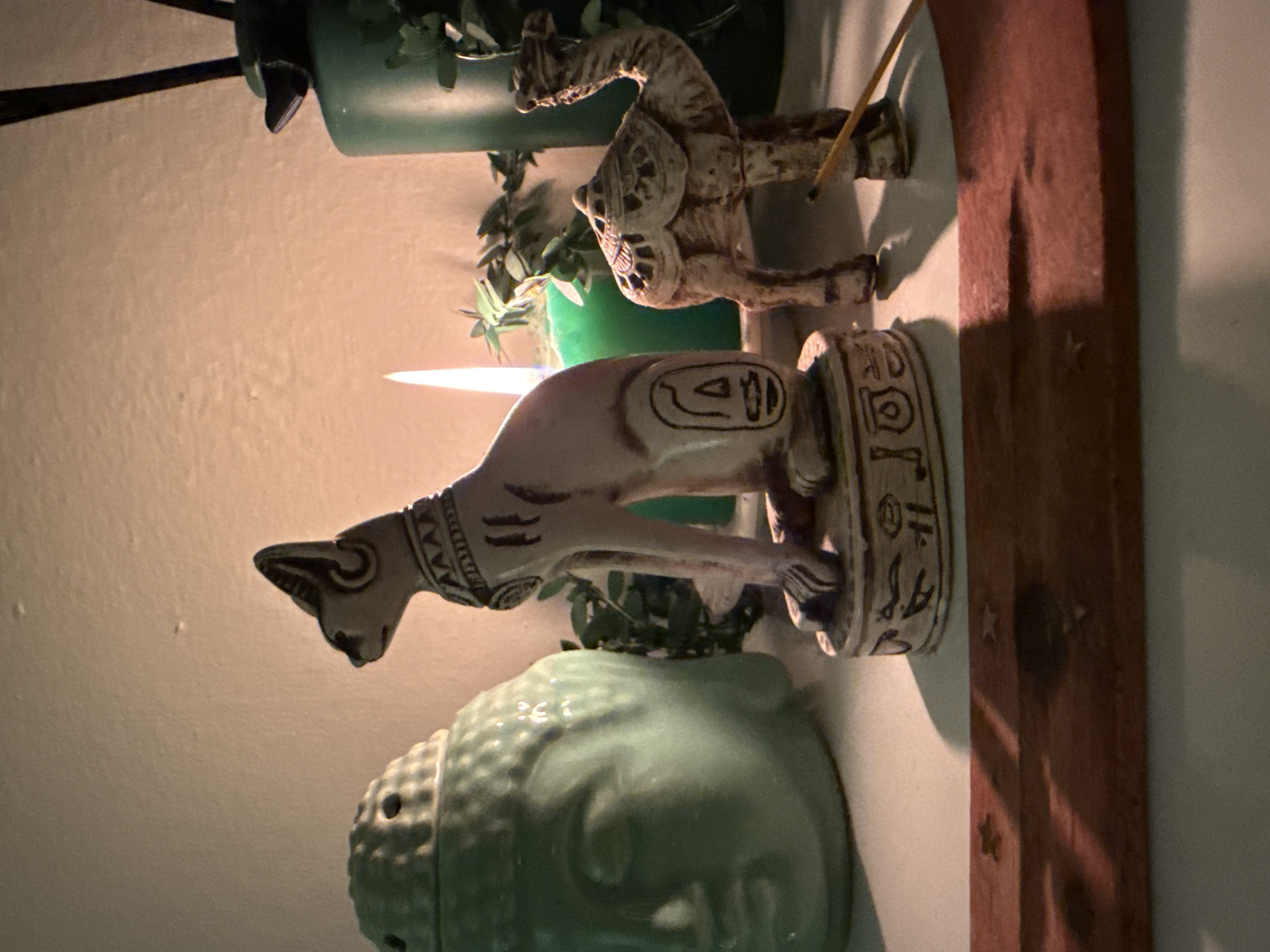 A close-up, eye-level shot shows a collection of decorative figurines on a white surface, possibly a shelf, illuminated by warm, soft lighting. In the foreground, a reddish-brown wooden incense holder with faint star imprints lies horizontally. Behind it, a light-colored, stylized figurine of a camel stands to the right, with an incense stick extending from its base. To the left of the camel is a statue of an Egyptian cat goddess, likely Bastet, with hieroglyphic markings on its base and collar. Further to the left, a large, pale green ceramic Buddha head, serving as an incense burner, is visible. A lit candle with a bright flame, possibly green, is positioned between the cat and Buddha statues, casting a glow. Behind the figurines, a tall, dark green vase with black reeds or sticks is partially visible on the right, alongside some green foliage. The background consists of a textured, off-white wall. The scene is indoors, with no discernible location in Kadıköy, Turkey.