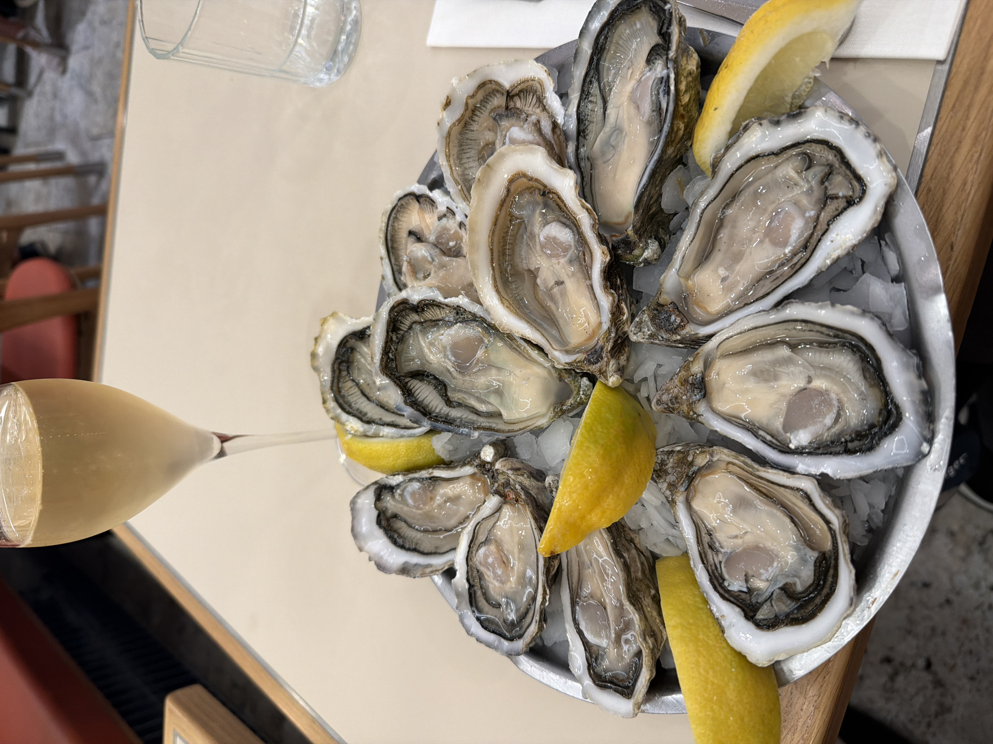 A high-angle shot captures a plate of fresh oysters served with lemon wedges atop crushed ice in a metal dish. A champagne flute rests horizontally to the left of the plate. The setting appears to be a restaurant, indicated by the table surface and blurred background elements. While the image doesn't explicitly confirm its location in Paris, France, the presence of oysters—a common dish in French cuisine—and the overall ambiance are consistent with typical dining experiences in Paris. No people are directly visible in the frame.