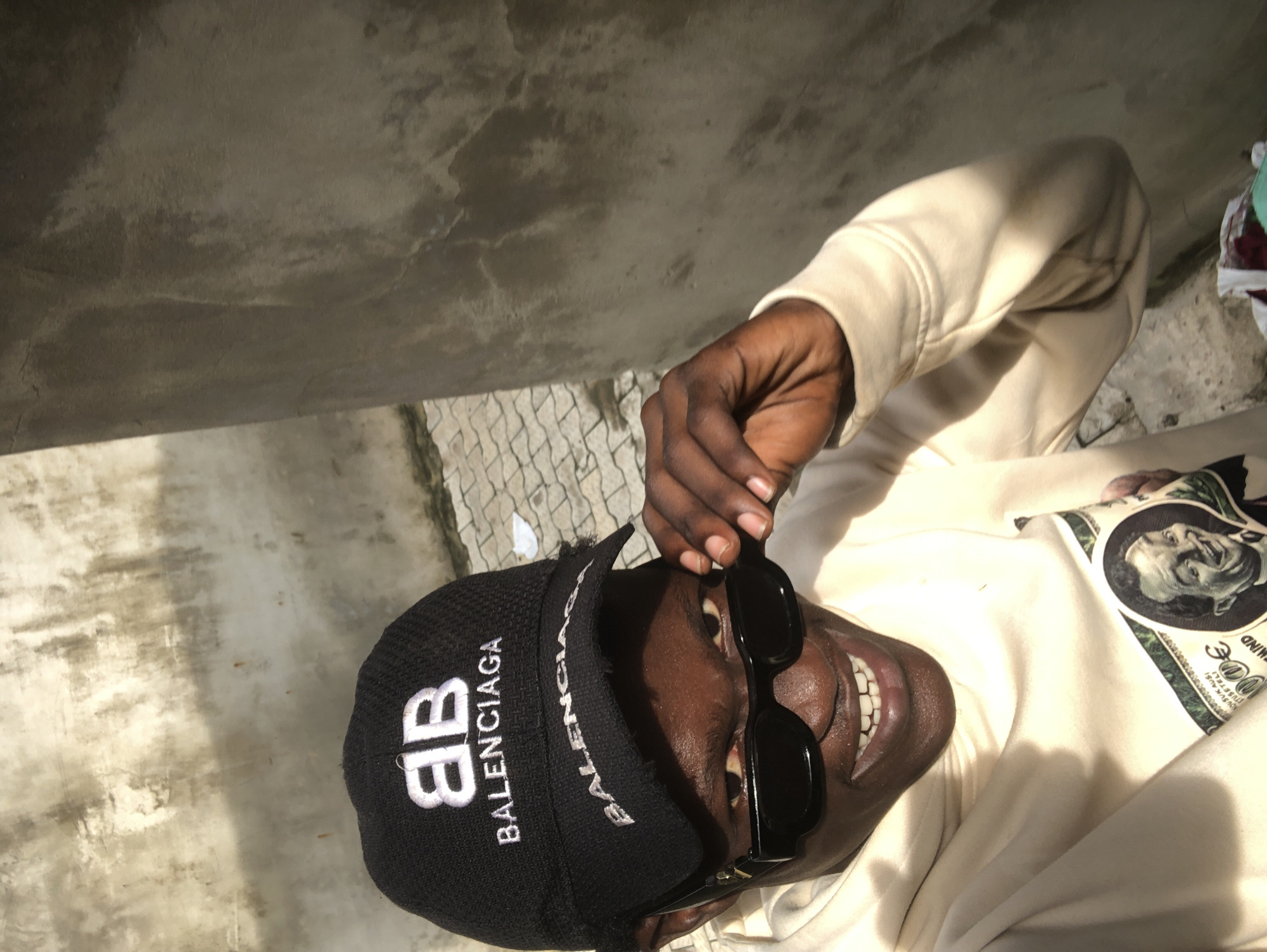 The image captures a close-up, upward-angled shot of a person, likely male, with dark skin, posing outdoors in Ado, Nigeria. The person is wearing a black baseball cap with "BALENCIAGA" written prominently in white, along with a "BB" logo. Black rectangular sunglasses rest on their face, and one hand, with dark skin, is seen holding the sunglasses slightly down, revealing their eyes. The person is looking upwards with a wide smile, showing white teeth. They are dressed in a light-colored, possibly beige or off-white, hooded sweatshirt. A graphic design is partially visible on the front of the sweatshirt, featuring a stylized image reminiscent of a dollar bill, including a portrait (possibly Benjamin Franklin) and text like "...100 DOLL..." and what appears to be "MIND". The background consists of a textured, light grey concrete wall or surface on the left, and a patch of light-colored, possibly tiled or patterned ground on the right. Strong sunlight illuminates the scene, casting distinct shadows and highlighting facial features and clothing details, suggesting it is daytime. The overall mood conveyed by the smile and direct gaze is positive and confident.