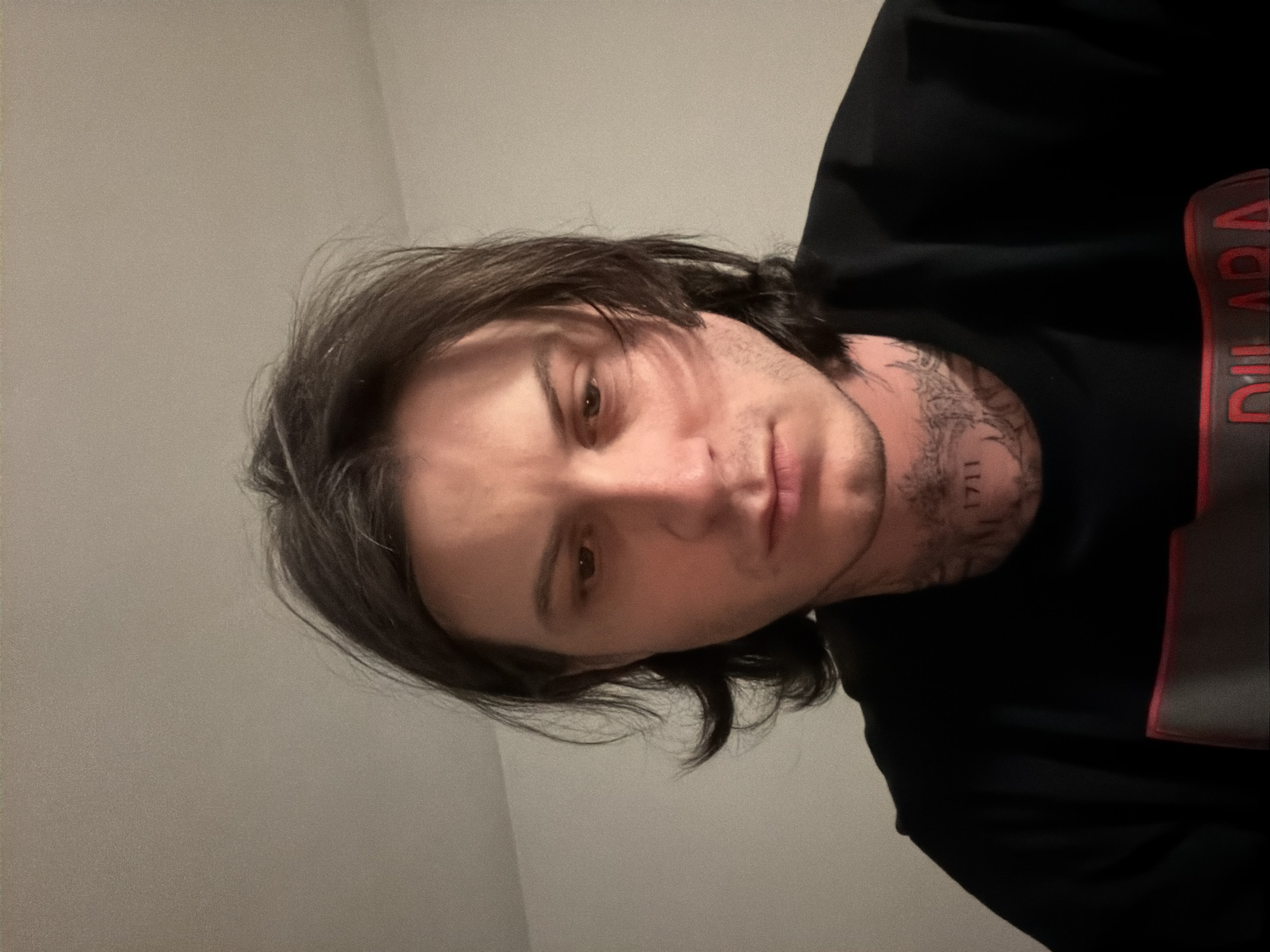 A portrait-style image, rotated 90 degrees counter-clockwise, depicts a male individual indoors. He has medium-length, dark brown hair styled casually, with some strands falling across his forehead. His face shows light stubble, and his expression is neutral, with his dark eyes looking towards the camera. Tattoos are visible on the right side of his neck (viewer's left), featuring intricate designs and what appears to be the number "1711".
He is wearing a dark, possibly black, long-sleeved top or sweatshirt. On the right side of his chest/shoulder area (viewer's left), there is a prominent red and dark grey design, with the word "DILARA" clearly visible in bold red letters.
The background is a plain, light grey or beige wall, suggesting an indoor setting with soft, even lighting. There are no distinguishing features in the background to indicate a specific location. The activity is a casual pose for a photograph, possibly a selfie. There are no visual cues within the image to confirm the provided context of London, United Kingdom. The image has a slightly grainy texture.