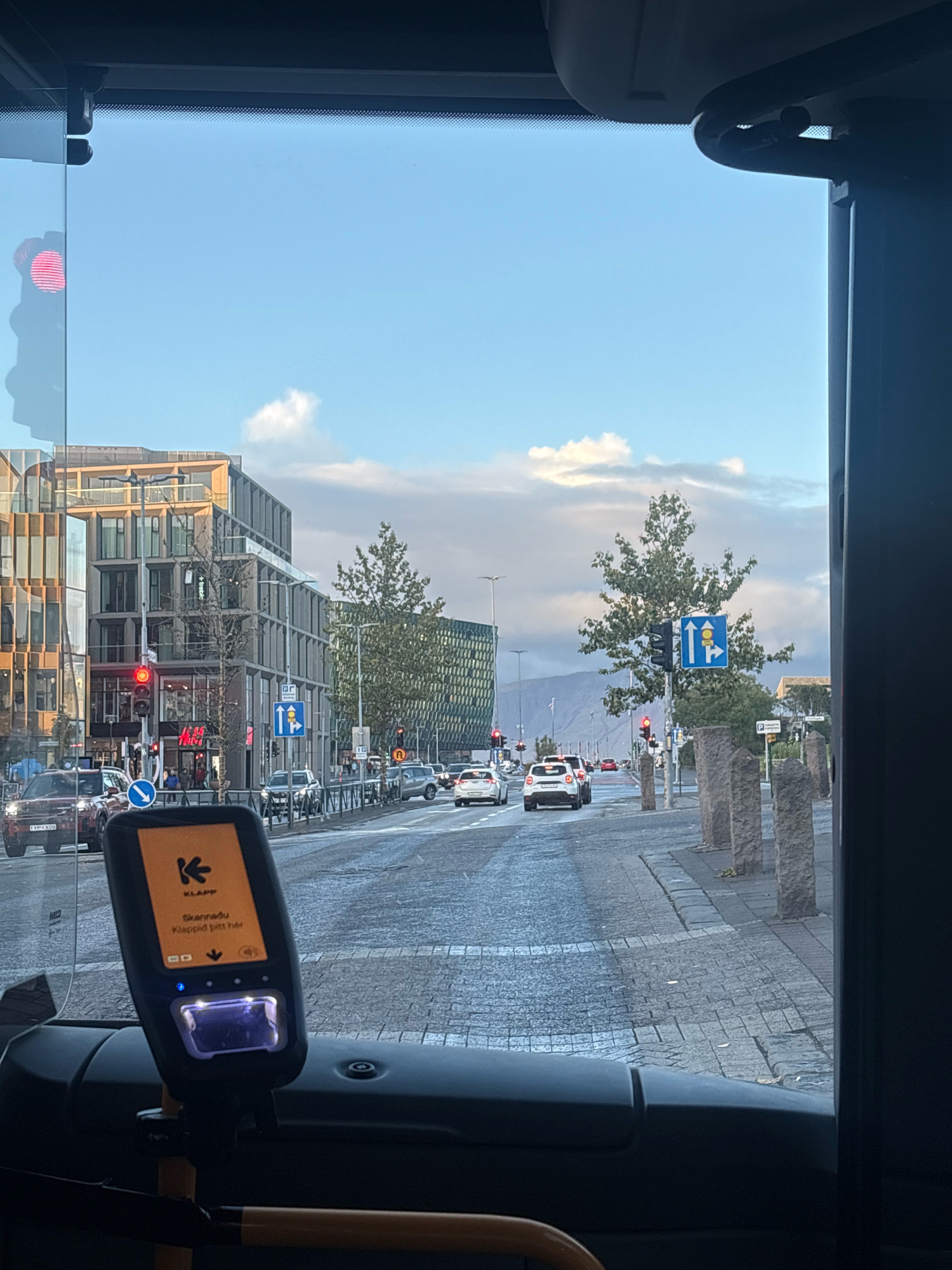 The image is a view from the driver's seat of a bus, looking out the windshield and side window at a street scene in Reykjavik, Iceland. In the foreground, a bus ticket validation machine is mounted on the dashboard. The machine has an orange screen with a logo and text in Icelandic, and a card reader slot. The street outside is paved with cobblestones and lined with stone pillars. There are several cars on the road, including a red SUV, a white hatchback, and other vehicles. Traffic lights are visible, with a red light illuminated on one. Buildings with modern architecture, some with glass facades, line the street. Trees are planted along the sidewalk, and a mountain range is visible in the distance under a cloudy sky. The overall scene depicts a typical urban environment in Reykjavik.
