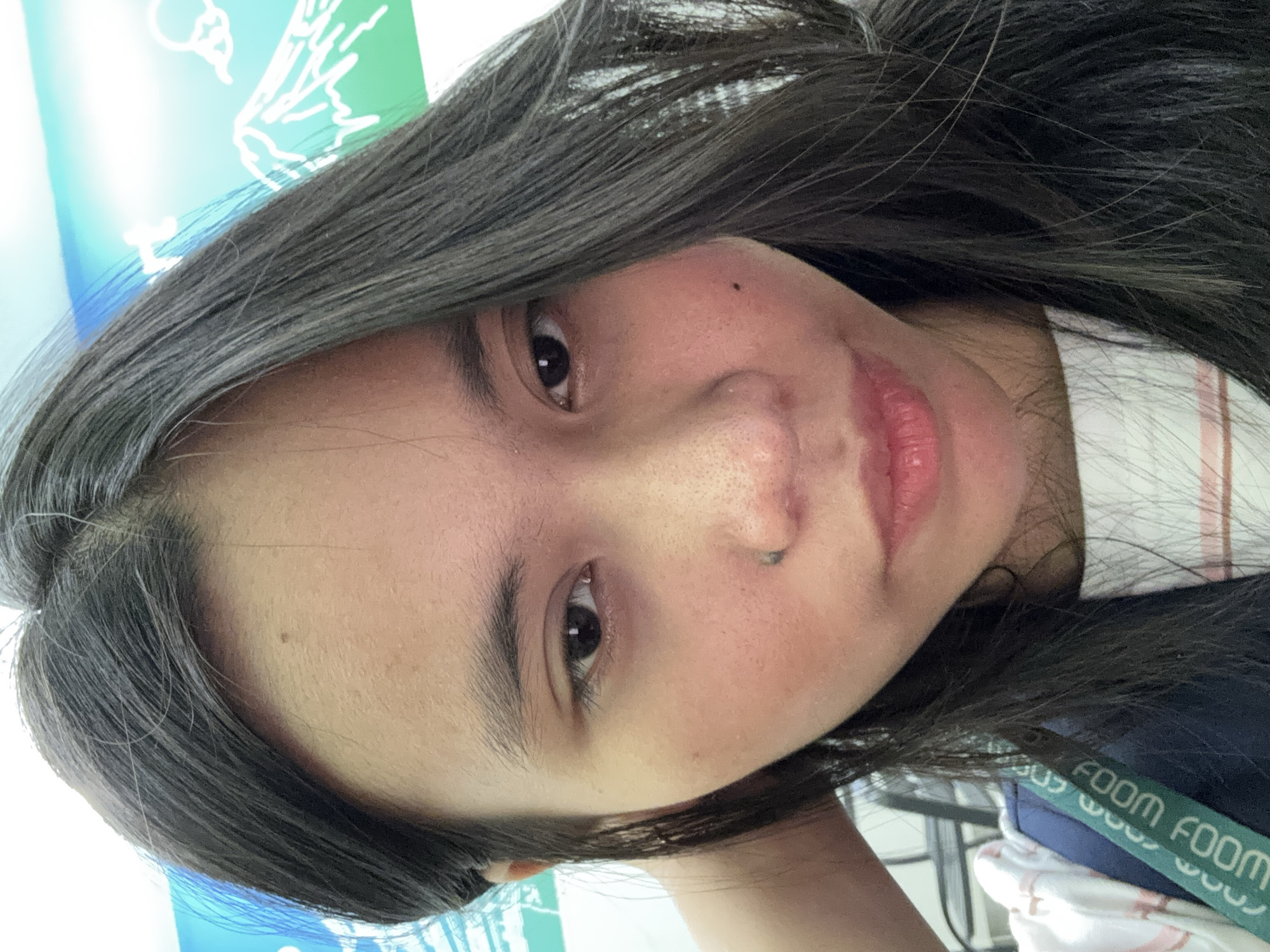 The image is a close-up, selfie-style portrait of a young woman with dark hair, taken indoors. She is looking directly at the camera. Her face is slightly turned to the left, and her right hand is raised to her head. She is wearing a white t-shirt with a pink stripe and a dark blue strap from a bag across her chest. The background appears to be a wall with a blue and green graphic of mountains. The lighting is bright, possibly from overhead fluorescent lights common in commercial settings. The specific location in East Jakarta, Indonesia, is not identifiable from the visual information provided.