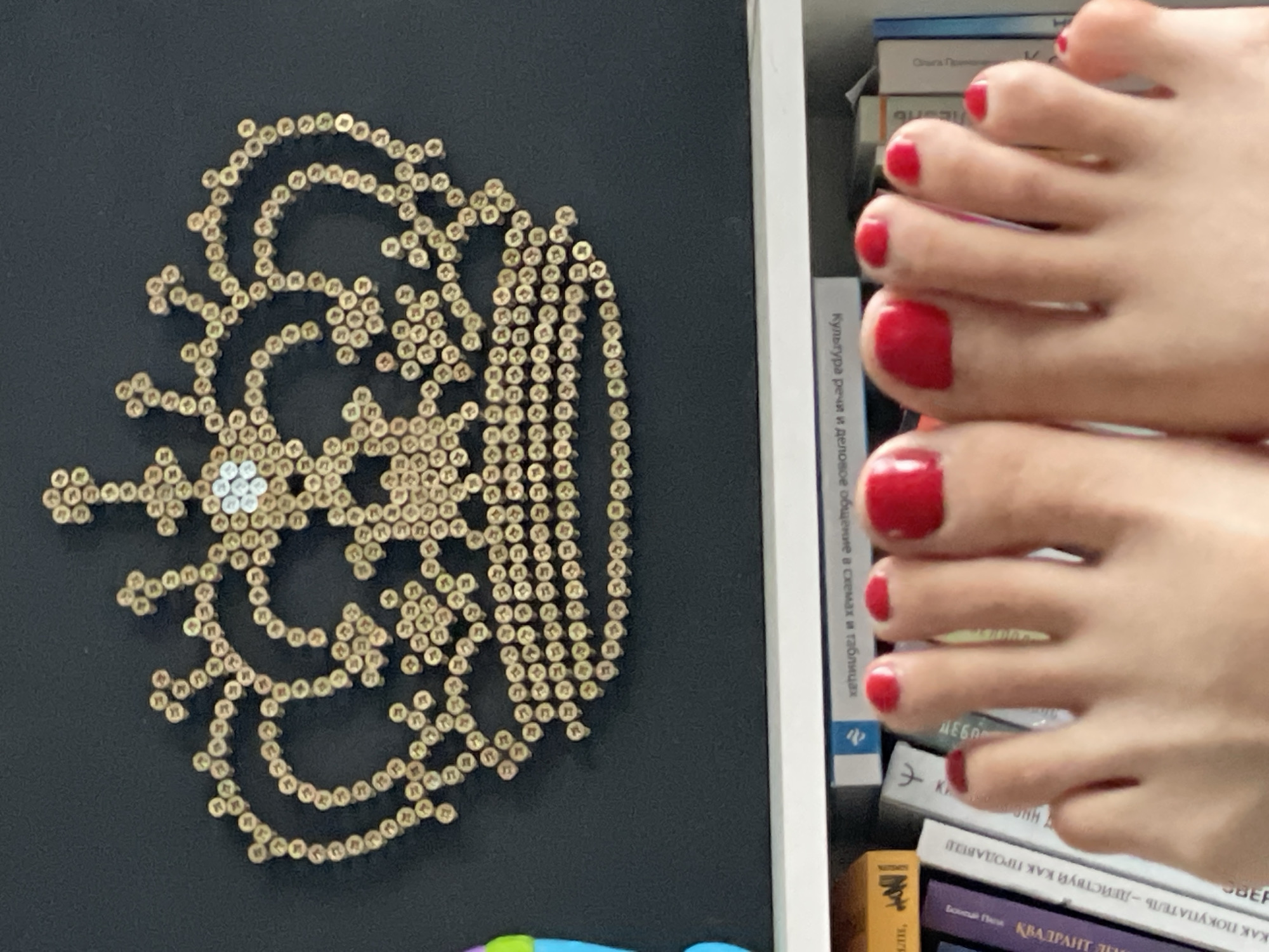 Here is a forensic description of the image:
The image shows a composition of disparate elements. On the left, there's a black surface displaying an ornate, lace-like design crafted from small, cylindrical objects, possibly beads or rivets, arranged in curved, swirling patterns. At the center of the design is a cluster of silver-colored elements.
To the right of this design is a bookshelf containing several books with titles in a language that appears to be Russian. Superimposed over the books are two bare feet with toes painted a bright red color. The feet are positioned such that the toes are visible against the backdrop of the book spines. The toenails are neatly manicured.
The feet appear to belong to a fair-skinned individual. The background is slightly blurred. The overall impression is a somewhat surreal juxtaposition of objects and body parts within an indoor environment.