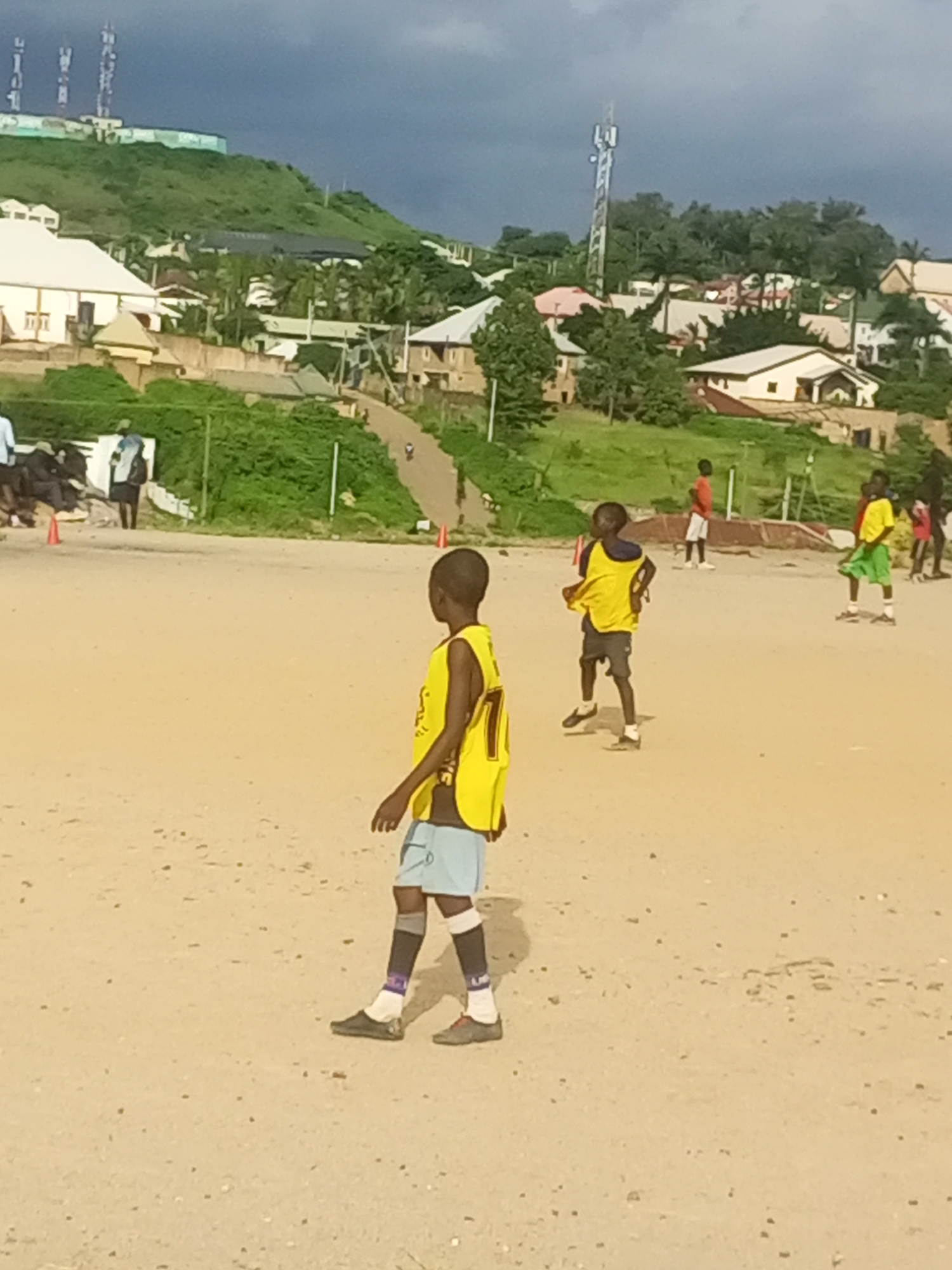 In Jos, Nigeria, a group of young boys are playing soccer on a dusty field. The scene is set against a backdrop of lush green hills dotted with houses and several tall communication towers. The sky is overcast with dark clouds, suggesting the possibility of rain.
The foreground features a boy in a yellow soccer jersey and light blue shorts, his back to the camera. He is wearing dark socks with purple bands and dark shoes. Further back, other boys are visible in athletic wear, some also in yellow jerseys, indicating they are on the same team or engaged in a game. One boy in a red shirt and white shorts stands further away, possibly a player on the opposing team or a spectator. The field is dry and sandy, with a few scattered pebbles. In the distance, a winding road leads up the hill, with small figures of people walking or driving. The buildings on the hill are a mix of styles, with white-tiled roofs and beige walls, characteristic of the region. The overall impression is of an active afternoon soccer game taking place in a semi-urban environment with a striking natural landscape. There is no visible text in the image.