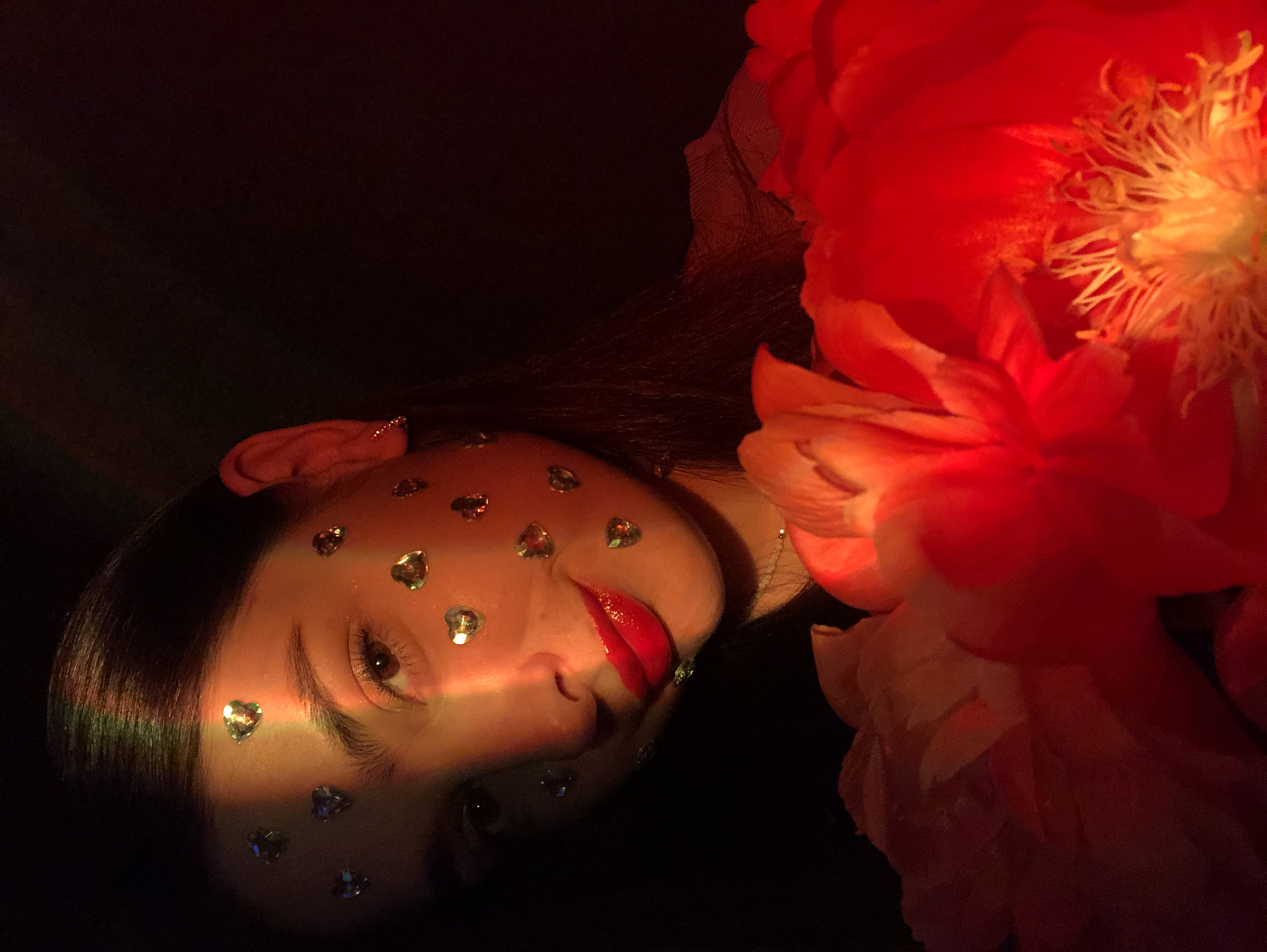 In Noviy, Russia, a young woman with dark hair and brown eyes is pictured in a dimly lit indoor setting. She has red lipstick on and several small, heart-shaped rhinestones affixed to her cheeks and forehead. A rainbow-colored light stripe illuminates her face diagonally from the upper left to the middle right. In the foreground, out of focus, are bright red flowers. The overall impression is of a portrait with dramatic lighting and decorative elements.