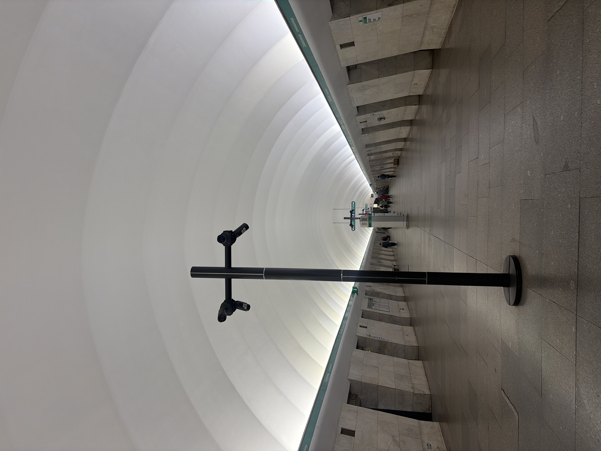 The image shows a platform in the Admiralteyskaya metro station in Saint Petersburg, Russia. The station has a distinctive architectural design with a large, arched ceiling.
A black metal structure with four security cameras is positioned in the foreground. The platform floor is paved with gray stone tiles. Along the walls are decorative architectural elements and recessed rectangular niches.
In the distance, a few people are waiting or walking on the platform. A station information kiosk is partially visible. The overall lighting is bright and diffused.