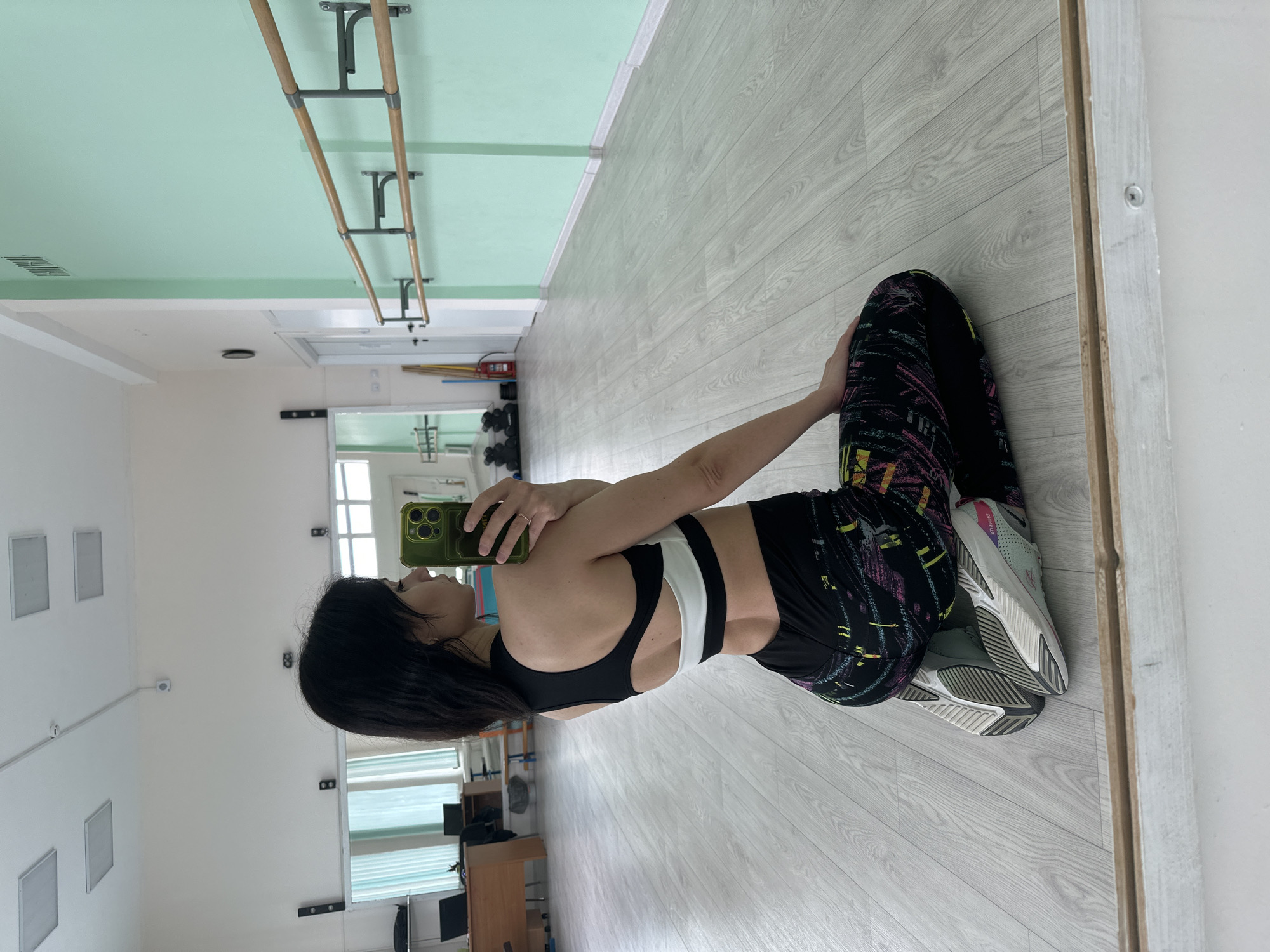 A woman is positioned in a room with a light green ceiling and light gray wood-patterned flooring, holding a cell phone in her right hand in front of her face, apparently taking a picture of her reflection in a mirror. She is wearing a black and white athletic bra and black leggings with a colorful geometric pattern, along with gray athletic shoes. The room appears to be a dance or exercise studio, as suggested by the barre mounted on the wall. A partially visible doorway can be seen in the background, leading to another room with office furniture. The exact location and city are not discernible from the image.